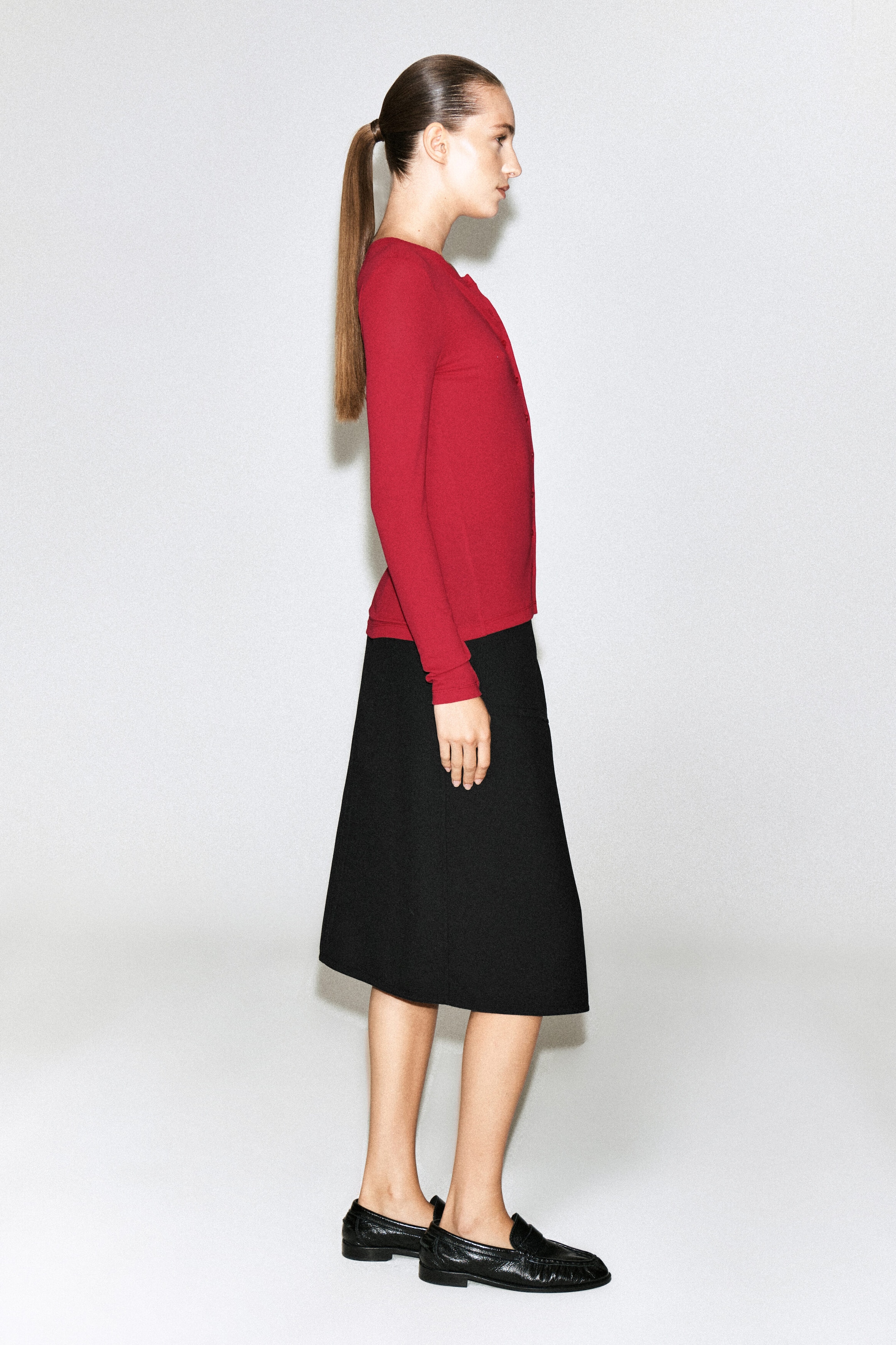 A red jersey cardigan top with long sleeves and a button-front closure is worn by the model. The top is styled with a black knee-length skirt and black loafers as the model stands in profile. A red jersey cardigan top with long sleeves and a button-front closure is worn by the model. The top is styled with a black knee-length skirt and black loafers as the model stands in profile.