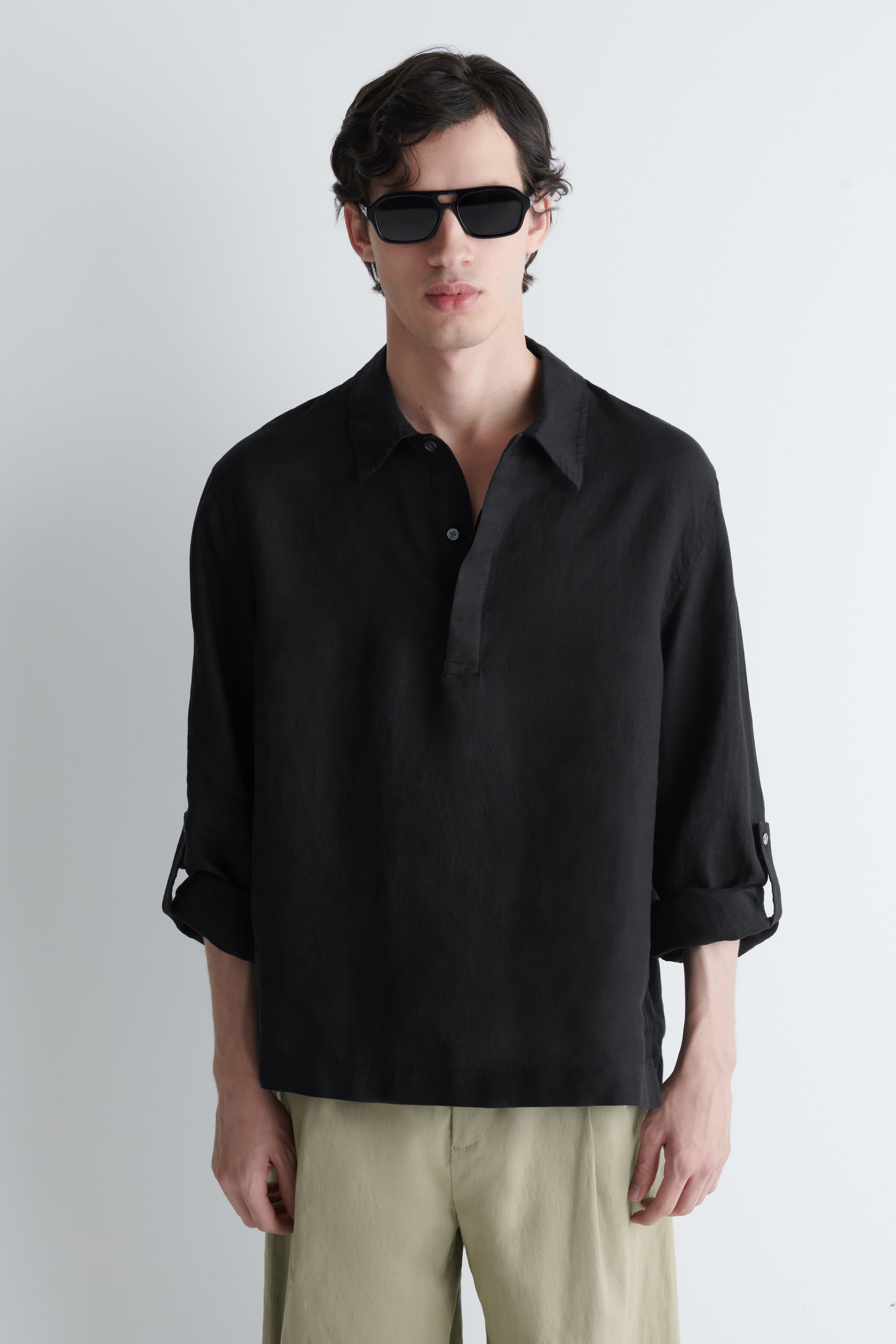 Visa större bild: En svart linneskjorta visas framifrån, med en avslappnad passform, en spridd krage och en halvknäppning med synliga knappar. Dess långa ärmar är upprullade och hålls på plats med knappflikar.