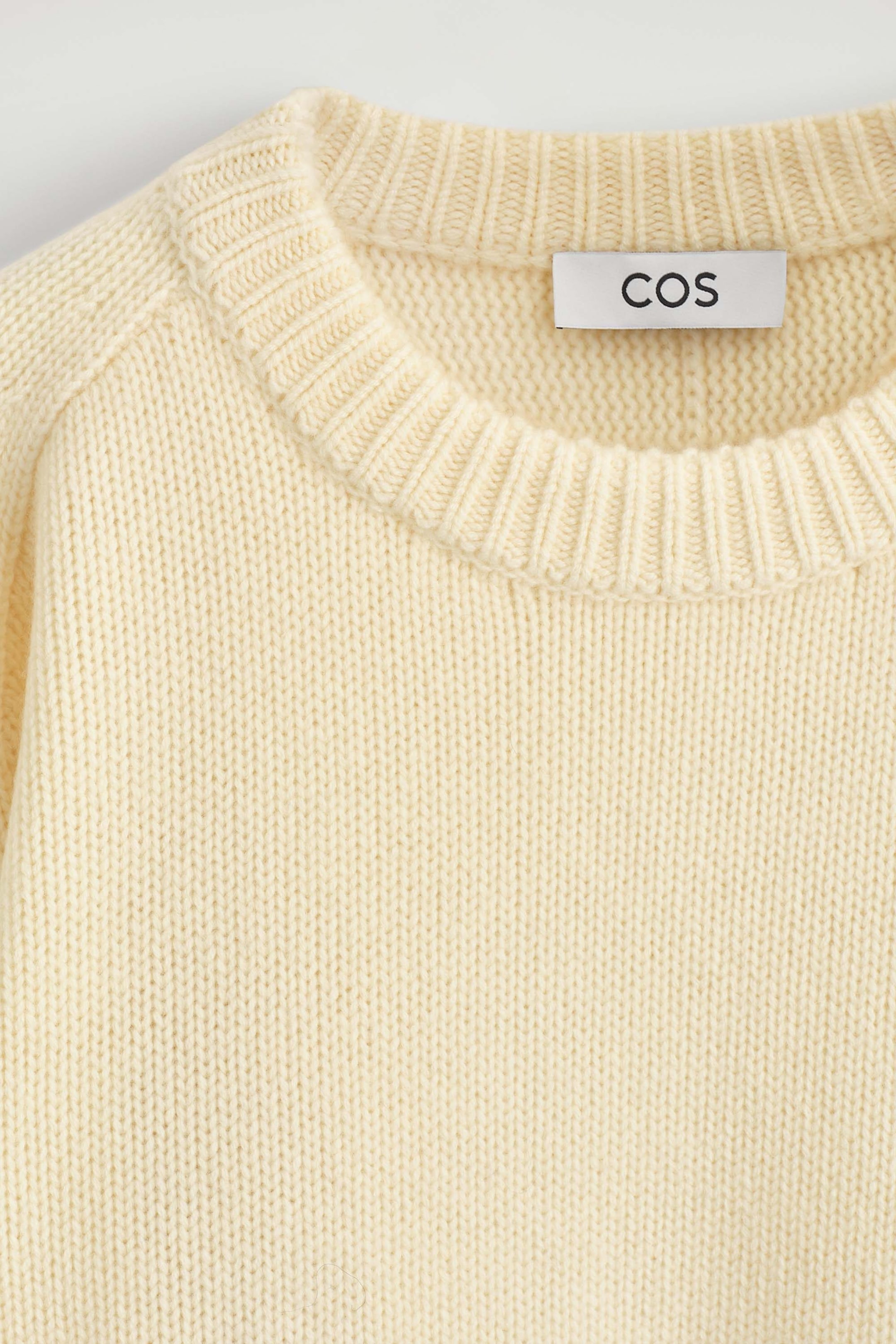 The rounded cashmere crew-neck jumper is shown in close-up, highlighting its cream-coloured, textured knit construction and thick ribbed neckline. The front-facing view highlights the raglan shoulder seams, with a white brand label reading "COS" visible on the inner collar. The rounded cashmere crew-neck jumper is shown in close-up, highlighting its cream-coloured, textured knit construction and thick ribbed neckline. The front-facing view highlights the raglan shoulder seams, with a white brand label reading "COS" visible on the inner collar.