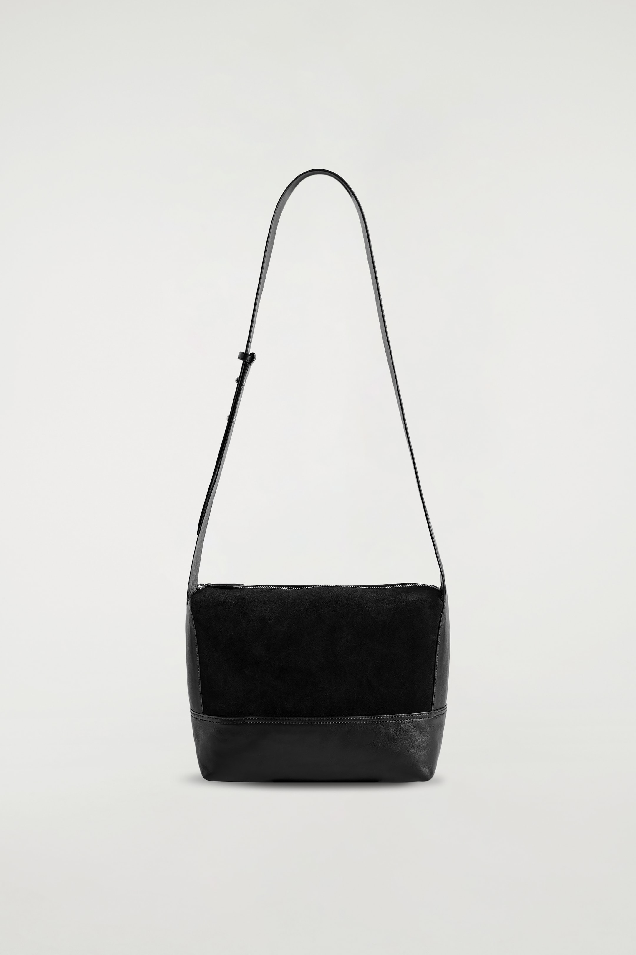 Agrandir l'image: Un sac à bandoulière noir est suspendu, présenté de face, avec un corps rectangulaire doté d'une section supérieure mate et texturée et d'une section inférieure en cuir lisse, divisées par une couture horizontale. Une fermeture éclair supérieure est visible ainsi qu'une fine sangle réglable.