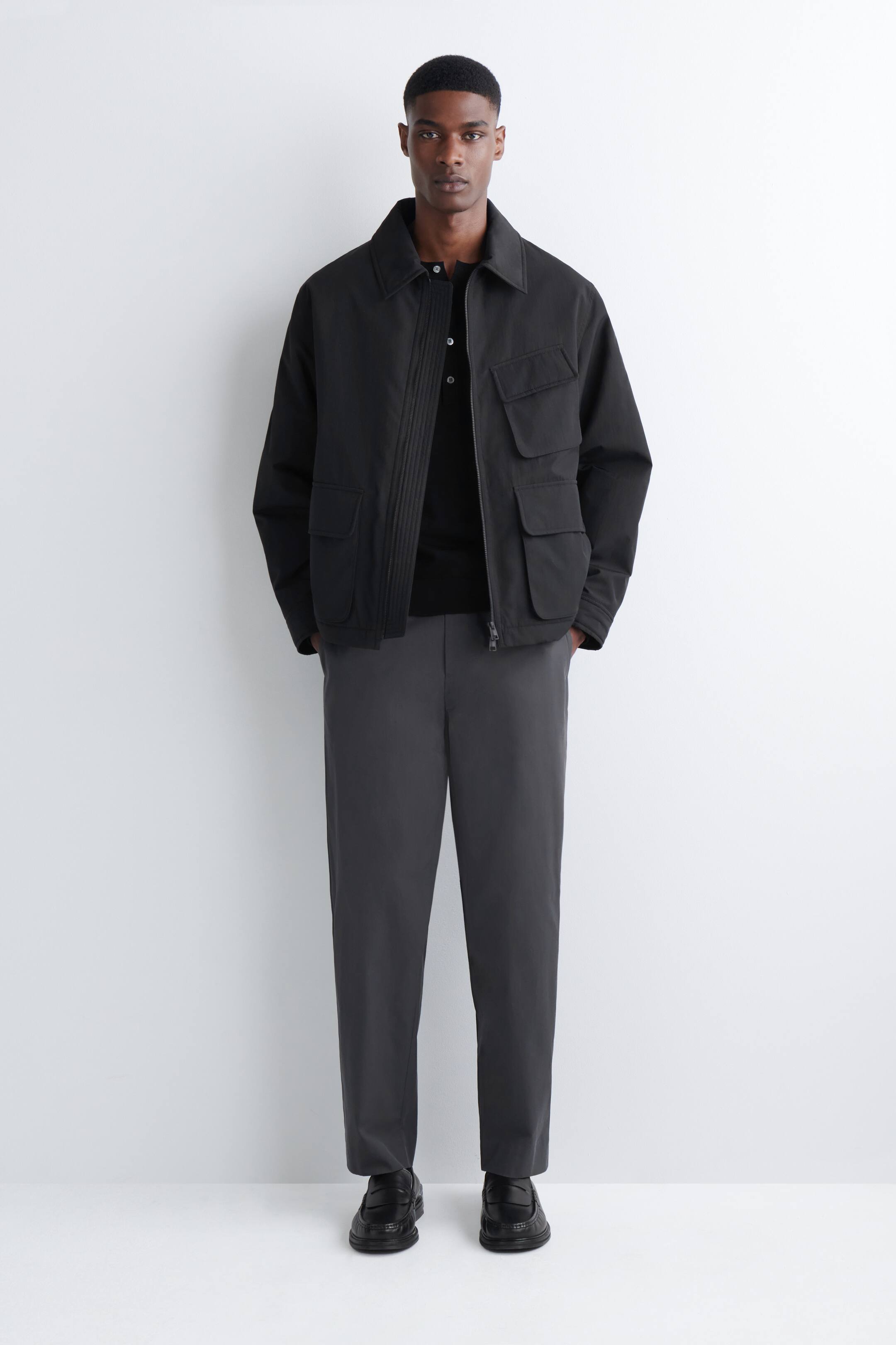 הצגת תמונה גדולה יותר: הדוגמן לובש מכנסי צ'ינו אפורים כהים עם גזרה ישרה. למכנסי הצ'ינו יש כיסים צדדיים נראים לעין והם משתפלים קלות מעל נעליים שחורות.