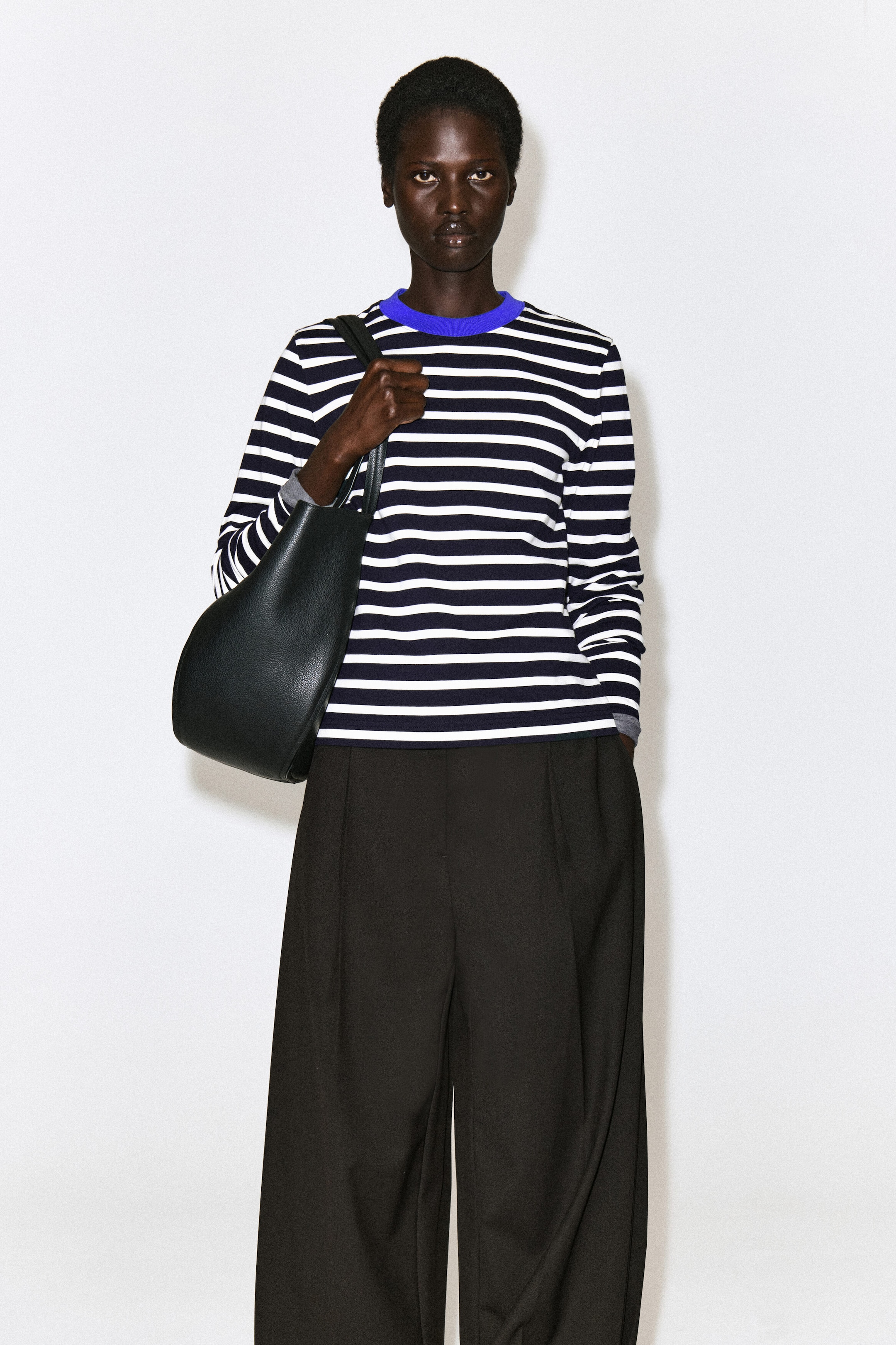 Ein schlicht geschnittenes Langarm-T-Shirt mit horizontalen Marineblau- und Weißstreifen sowie einem kräftig blauen Rundhalsausschnitt wird vom Model getragen. Das T-Shirt ist mit dunklen Wide-Leg-Hosen kombiniert, und eine schwarze Ledertasche wird über der Schulter getragen. Ein schlicht geschnittenes Langarm-T-Shirt mit horizontalen Marineblau- und Weißstreifen sowie einem kräftig blauen Rundhalsausschnitt wird vom Model getragen. Das T-Shirt ist mit dunklen Wide-Leg-Hosen kombiniert, und eine schwarze Ledertasche wird über der Schulter getragen.