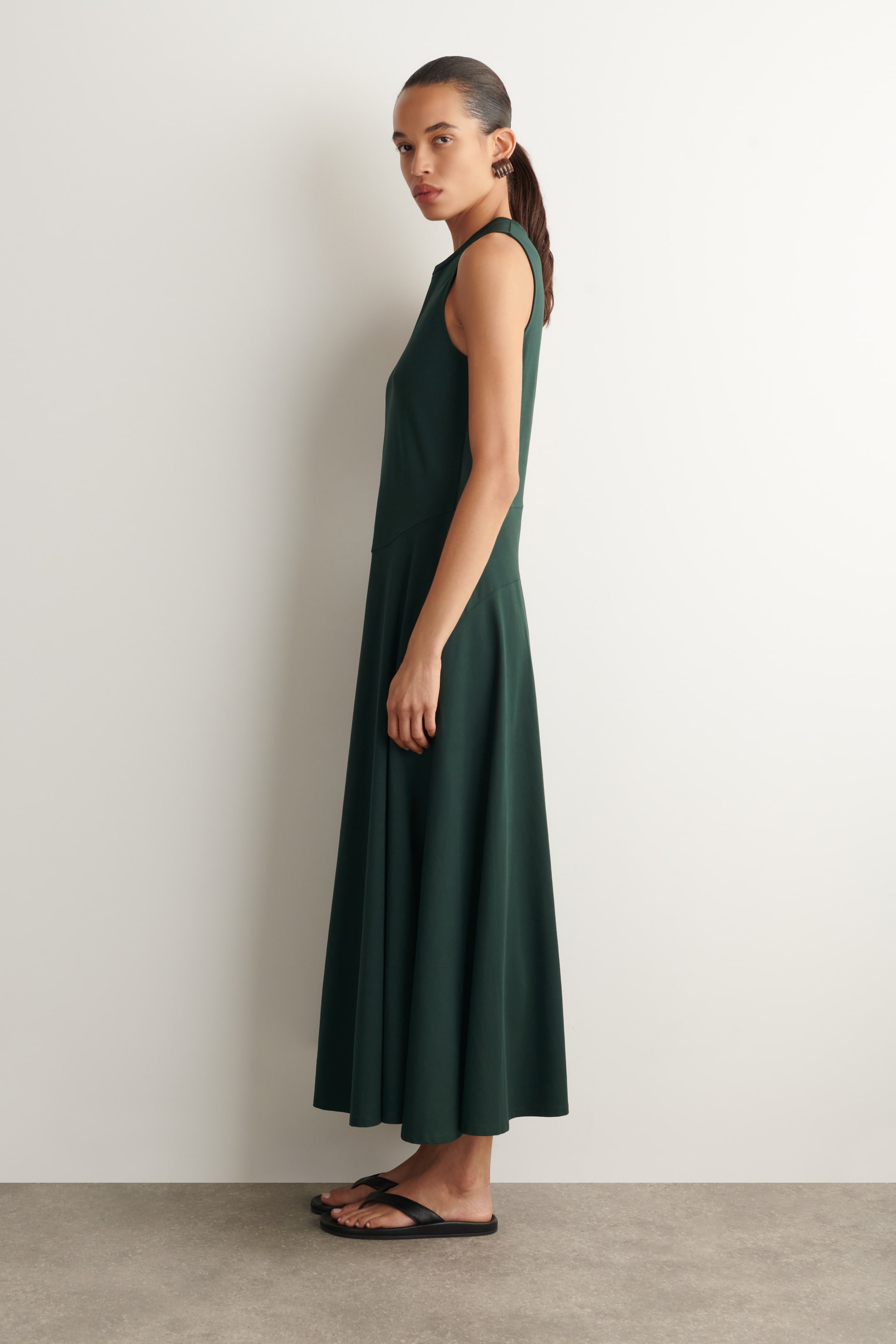 Grösseres Bild ansehen: Ein waldgrünes skulpturales Midikleid wird von der linken Seite gezeigt. Es zeichnet sich durch ein ärmelloses Design, geschwungene Nähte und einen sanft ausgestellten Midisaum aus.