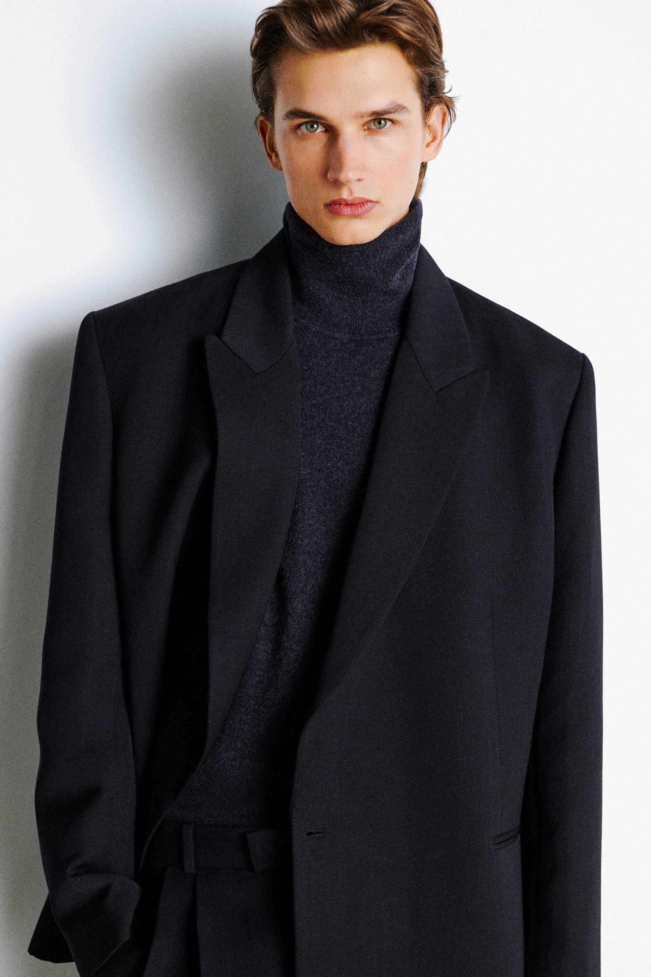 Un blazer croisé noir en laine mélangée, doté d’épaules structurées et de revers en pointe, est porté de face, superposé à un pull à col roulé en maille gris foncé. Le blazer à coupe décontractée est laissé ouvert, mettant en valeur la texture souple et le col montant du vêtement en maille en dessous. Un blazer croisé noir en laine mélangée, doté d’épaules structurées et de revers en pointe, est porté de face, superposé à un pull à col roulé en maille gris foncé. Le blazer à coupe décontractée est laissé ouvert, mettant en valeur la texture souple et le col montant du vêtement en maille en dessous.