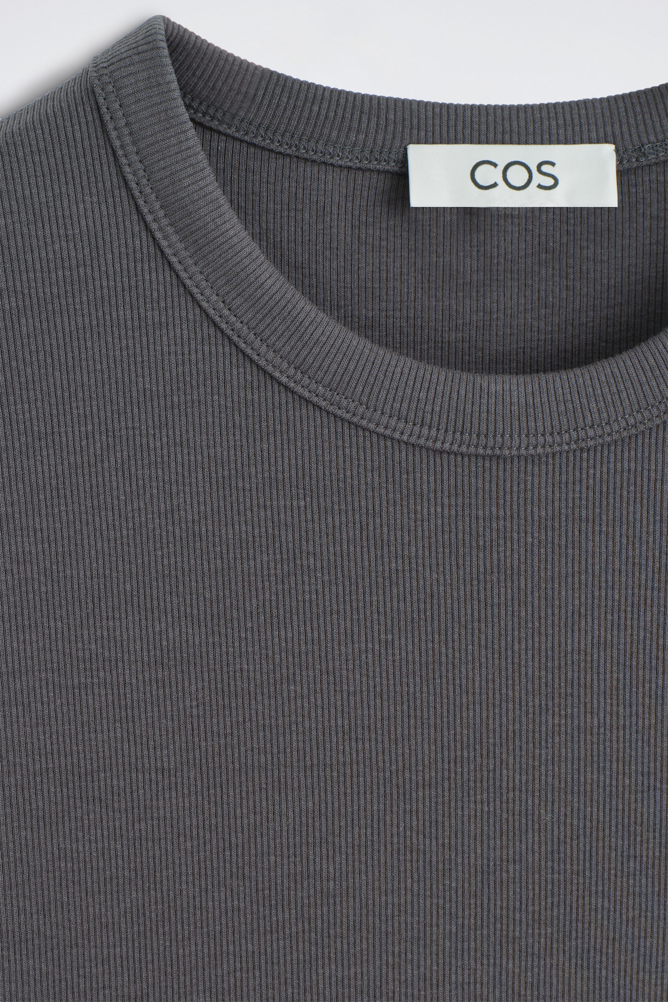 The slim ribbed cotton T-shirt in dark grey is showcasing its fine vertical rib-knit texture and crew neckline with double-stitched detailing. A white brand label reading "COS" is appearing on the inside back of the collar.