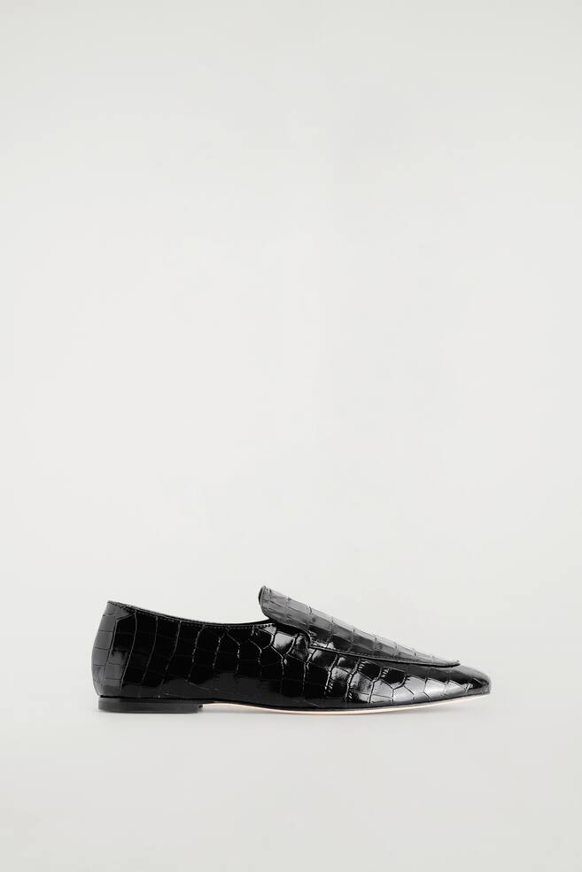 Een zwarte loafer van lakleer met een croc-effectstructuur is in zijaanzicht te zien, waarbij de glanzende afwerking en gestroomlijnde vorm worden benadrukt. De schoen heeft een hoge wreef met een opgestikte apron-naad en een lage, platte hak. - 1