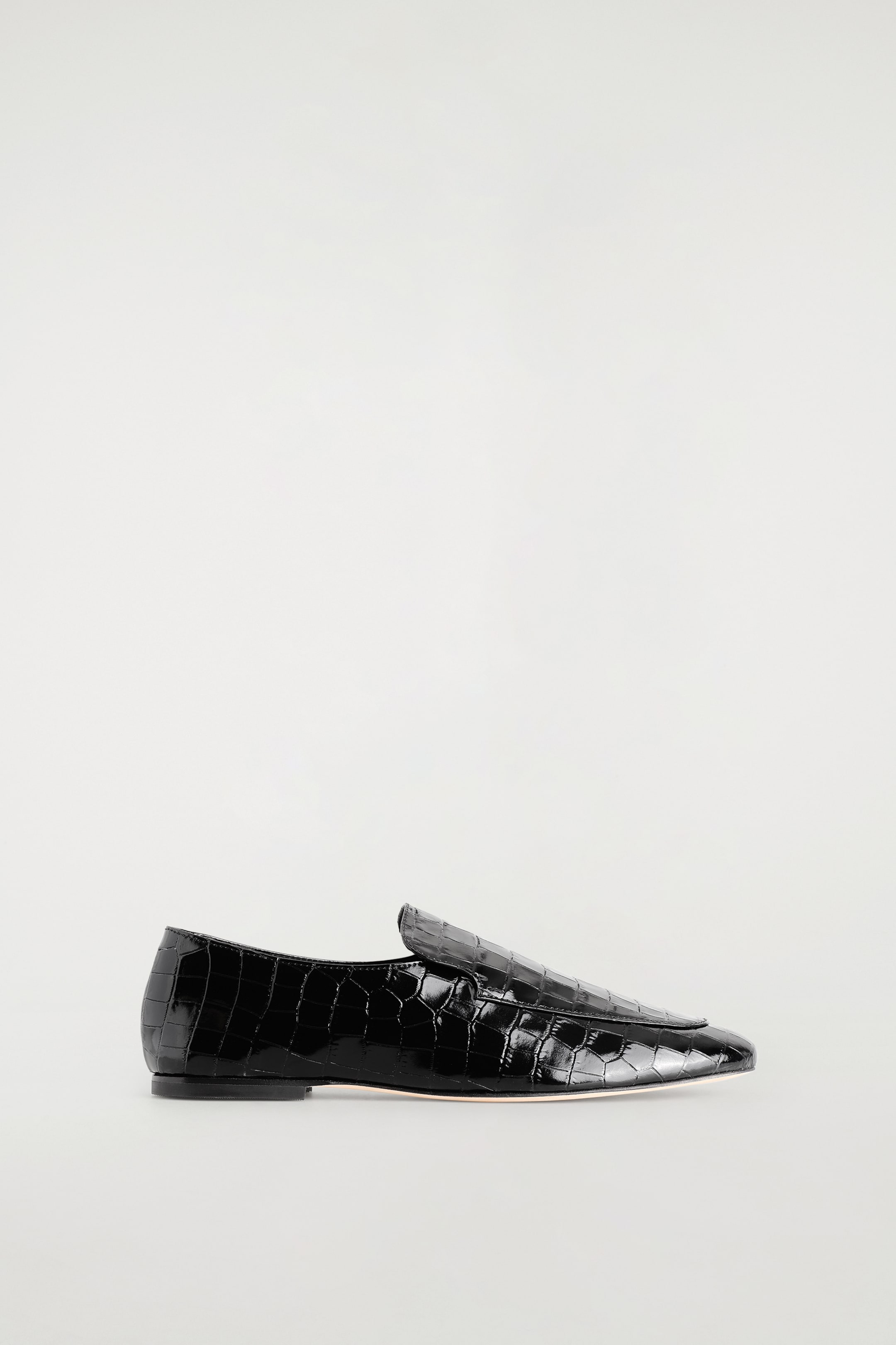 Größeres Bild ansehen: Ein schwarzer Loafer aus Lackleder mit Krokodilprägung ist in Seitenansicht zu sehen und präsentiert sein glänzendes Finish sowie seine schlanke Form. Der Schuh zeigt einen hohen Spann mit einer oberseitig abgesteppten Vorderkappe und einen niedrigen, flachen Absatz.
