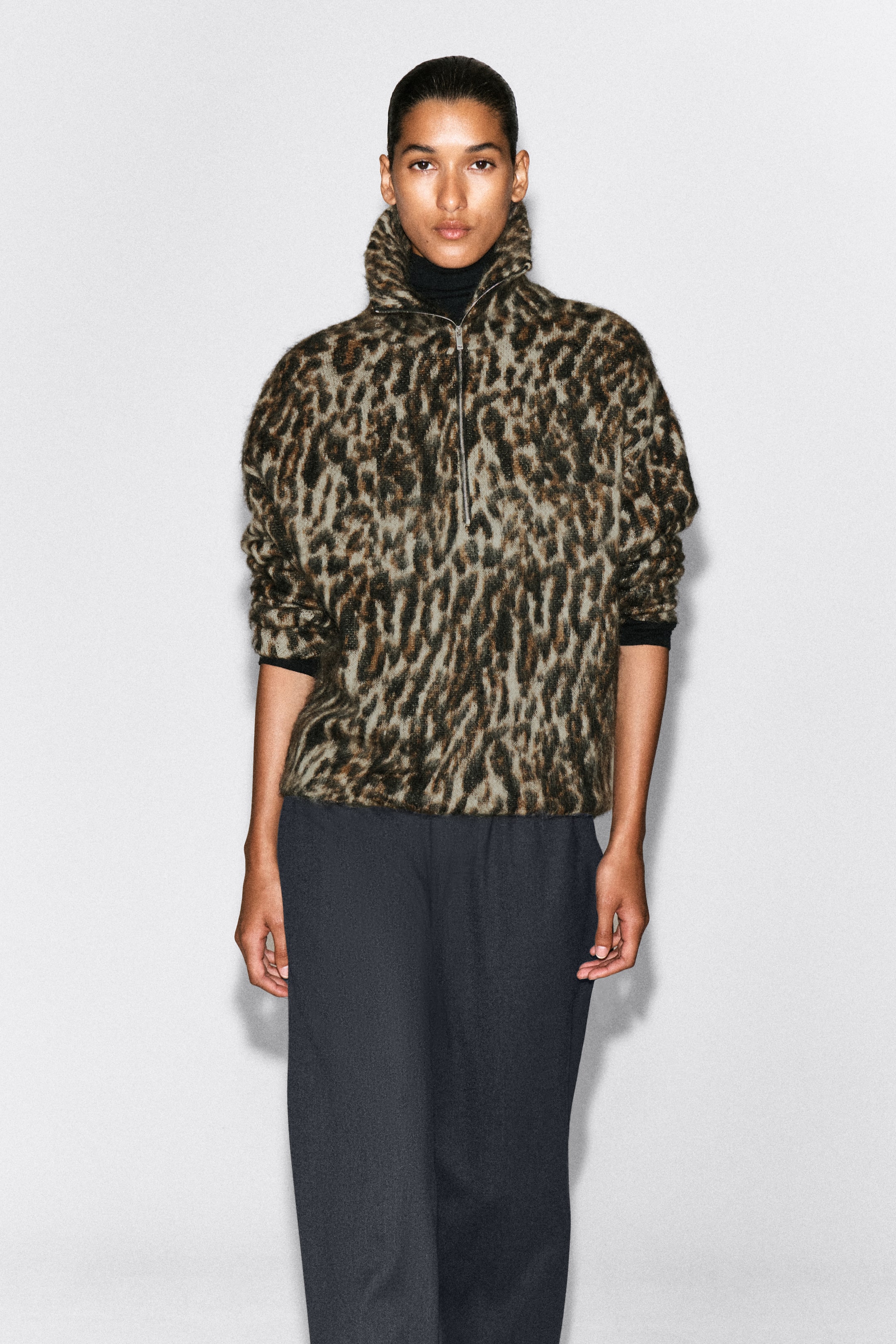 View larger image: A leopard-patterned brushed knit jumper is shown from the front, featuring a partially unzipped funnel neck. The jumper is worn layered over a black roll-neck and paired with dark trousers.