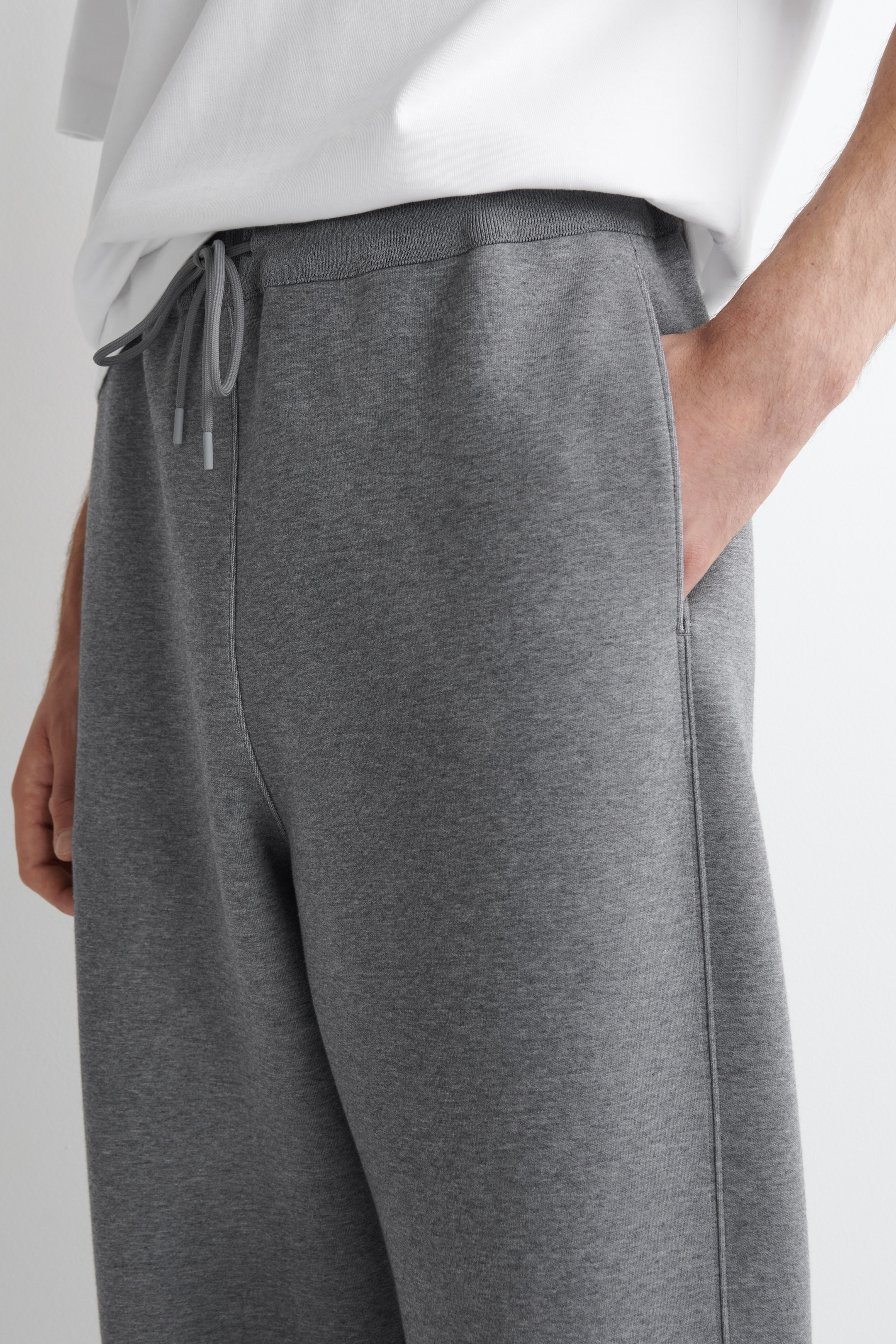 Größeres Bild ansehen: Graue Melange-Jogginghosen werden aus einem leichten Seitenwinkel präsentiert, mit einem gerippten elastischen Bund und einem gebundenen grauen Kordelzug. Eine Hand ruht in einer Seitentasche, eine markante vertikale Naht verläuft entlang des vorderen Beins, und ein weißes T-Shirt ist über dem Bund sichtbar.
