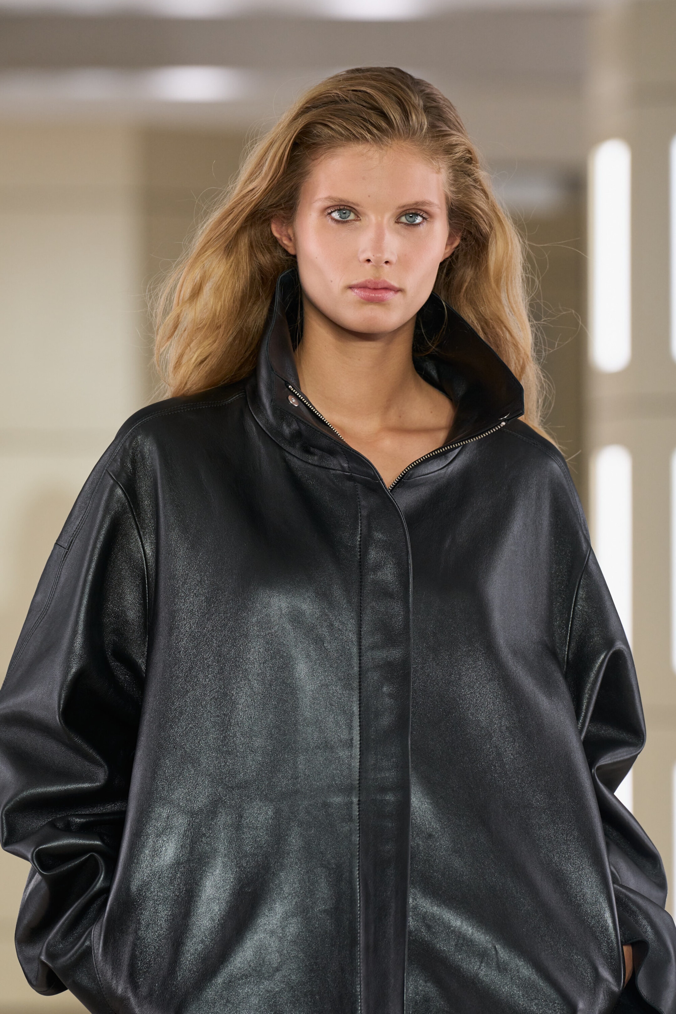 A black leather funnel-neck jacket is shown from the front, highlighting an oversized silhouette with dropped shoulders and a smooth, semi-matte finish. The jacket features a high, structured funnel collar that stands open at the neck, revealing a partially unzipped front closure concealed behind a clean placket. A black leather funnel-neck jacket is shown from the front, highlighting an oversized silhouette with dropped shoulders and a smooth, semi-matte finish. The jacket features a high, structured funnel collar that stands open at the neck, revealing a partially unzipped front closure concealed behind a clean placket.
