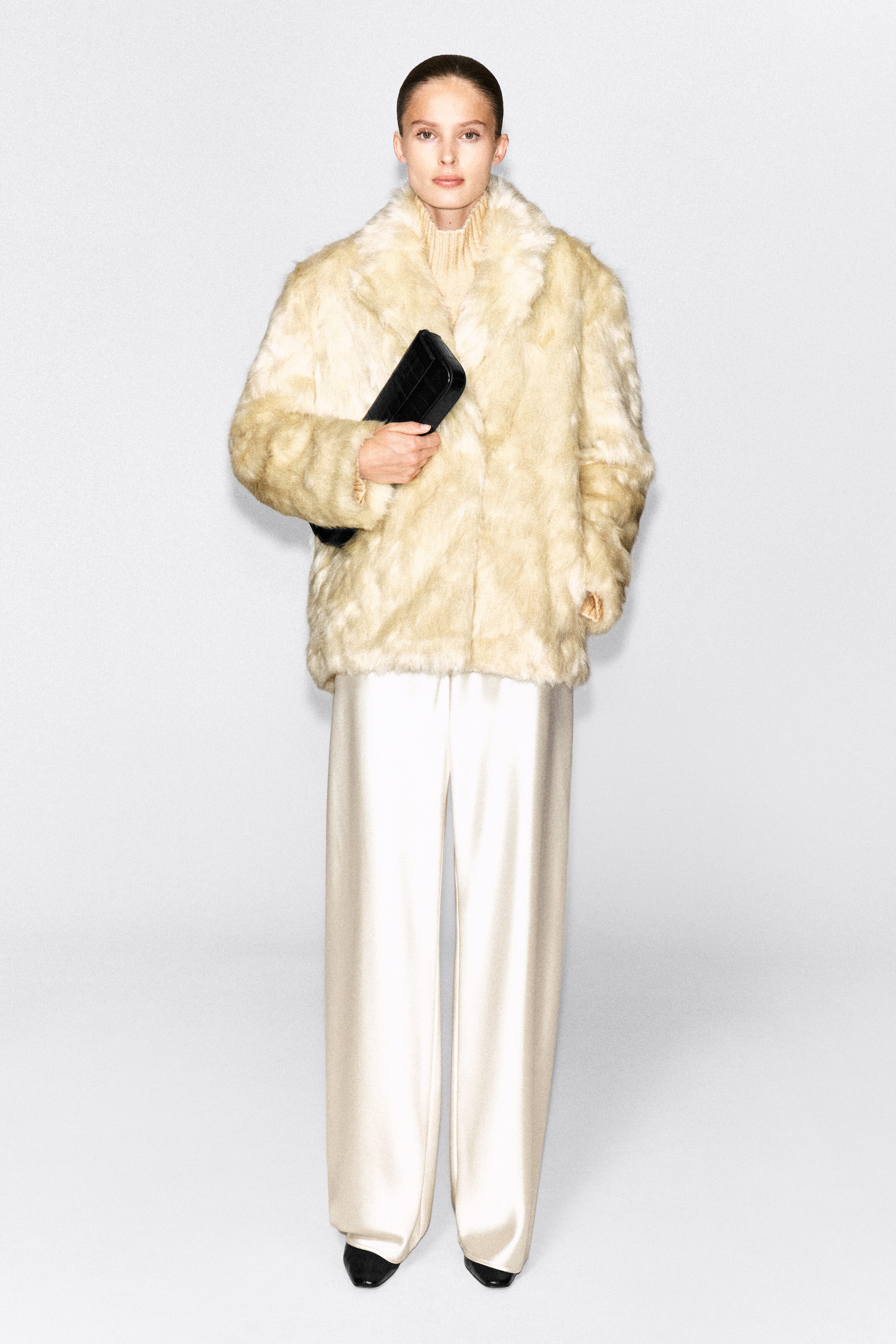 A cream-colored faux-fur jacket with notched lapels is worn by the model over a high-neck knit top and wide-leg satin trousers. The model is holding a black clutch bag in front of the jacket.
