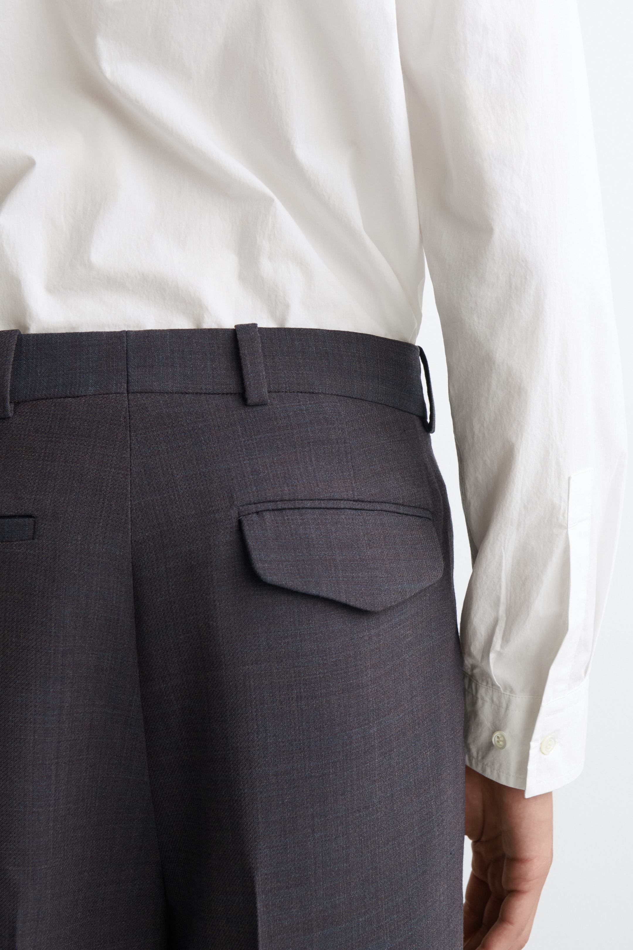 Agrandir l'image: Un pantalon gris foncé est vu de dos, avec une poche arrière à rabat pointu sur le côté droit et des passants de ceinture visibles le long de la ceinture. Une chemise blanche à manches longues est portée rentrée dans le pantalon.