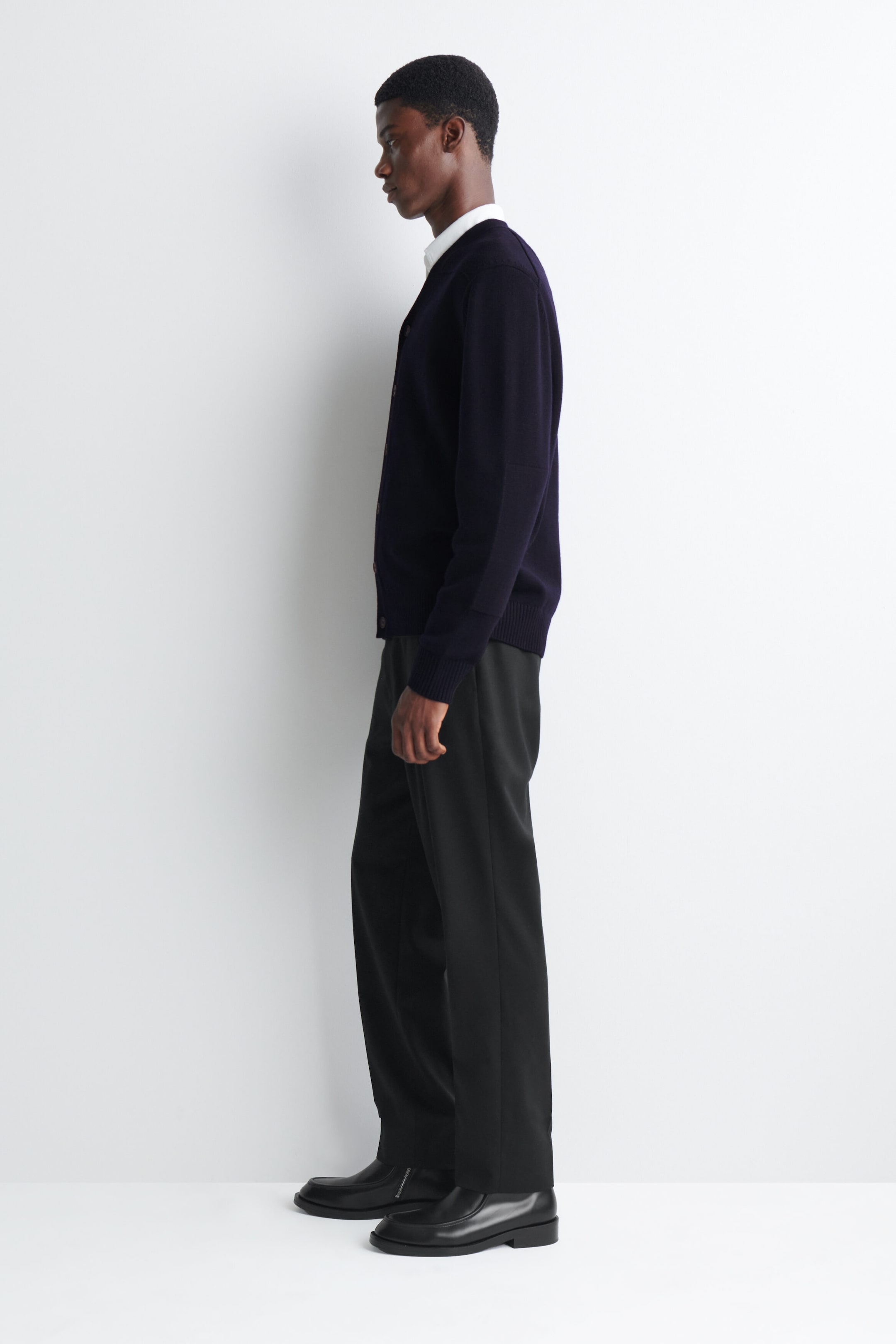 A dark navy blue knitted cardigan is being worn by the model, featuring a V-neck, button closures, and ribbed cuffs. The garment is layered over a white collared shirt and paired with black trousers and black leather boots. A dark navy blue knitted cardigan is being worn by the model, featuring a V-neck, button closures, and ribbed cuffs. The garment is layered over a white collared shirt and paired with black trousers and black leather boots.