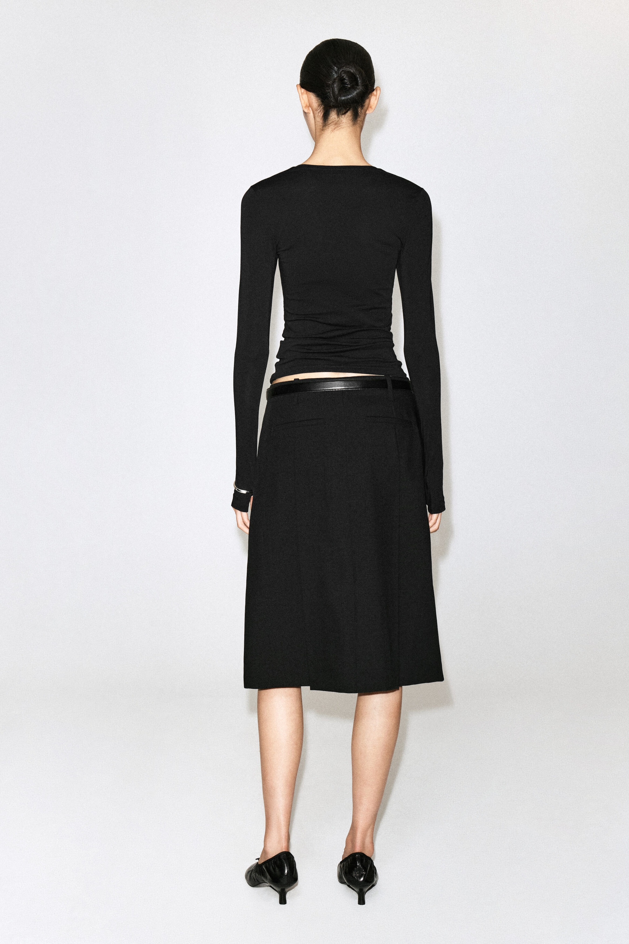 La mannequin porte une jupe noire longueur genou, associée à un haut noir ajusté à manches longues et à une fine ceinture en cuir noir. Elle se tient dos à l’appareil photo, mettant en valeur la silhouette épurée de la tenue, complétée par des escarpins noirs à petit talon.