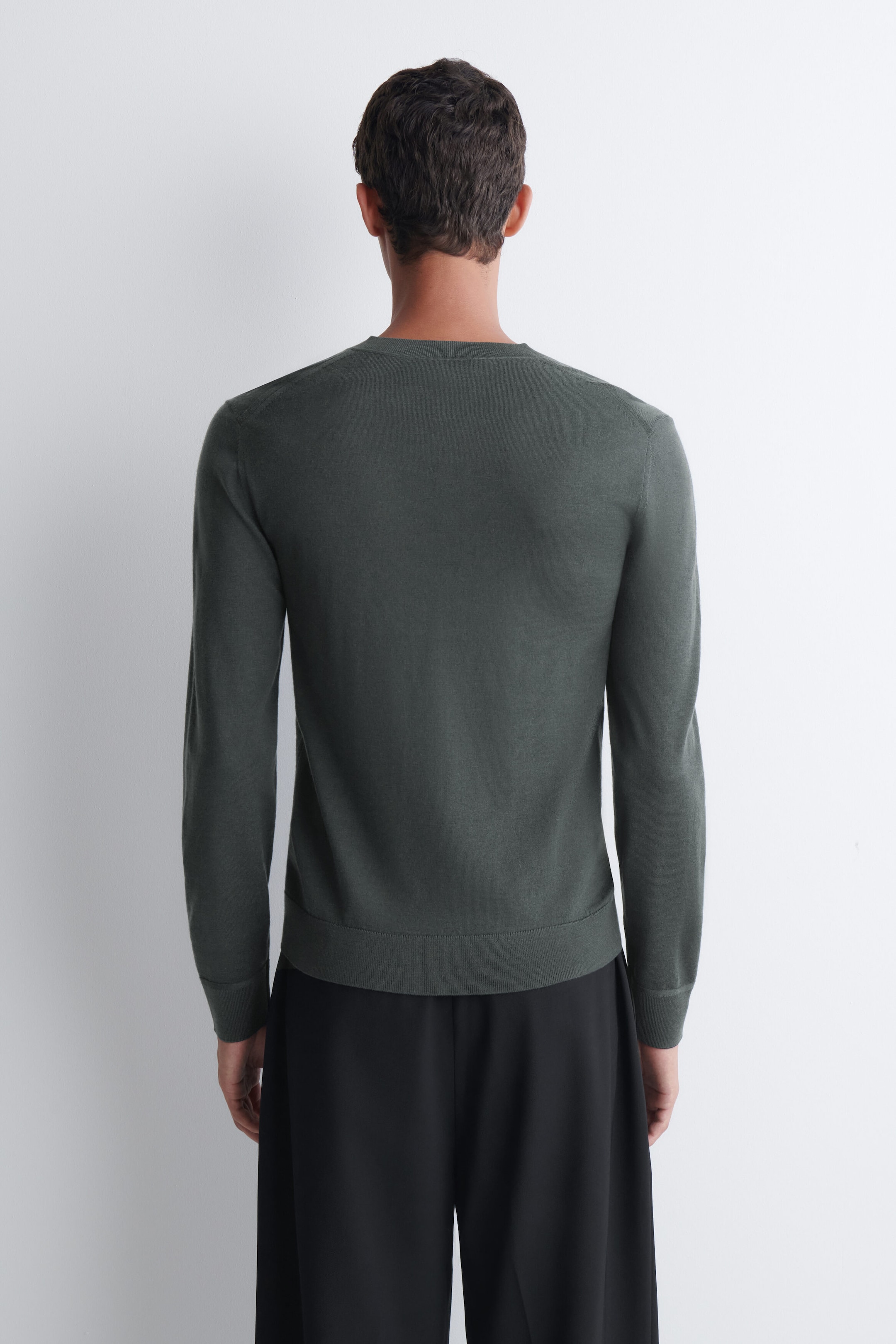 A slim merino wool Henley top in dark green is being worn by the model, shown from behind. The long-sleeve, fine-knit top features a ribbed hem and ribbed cuffs, and is styled with black trousers.