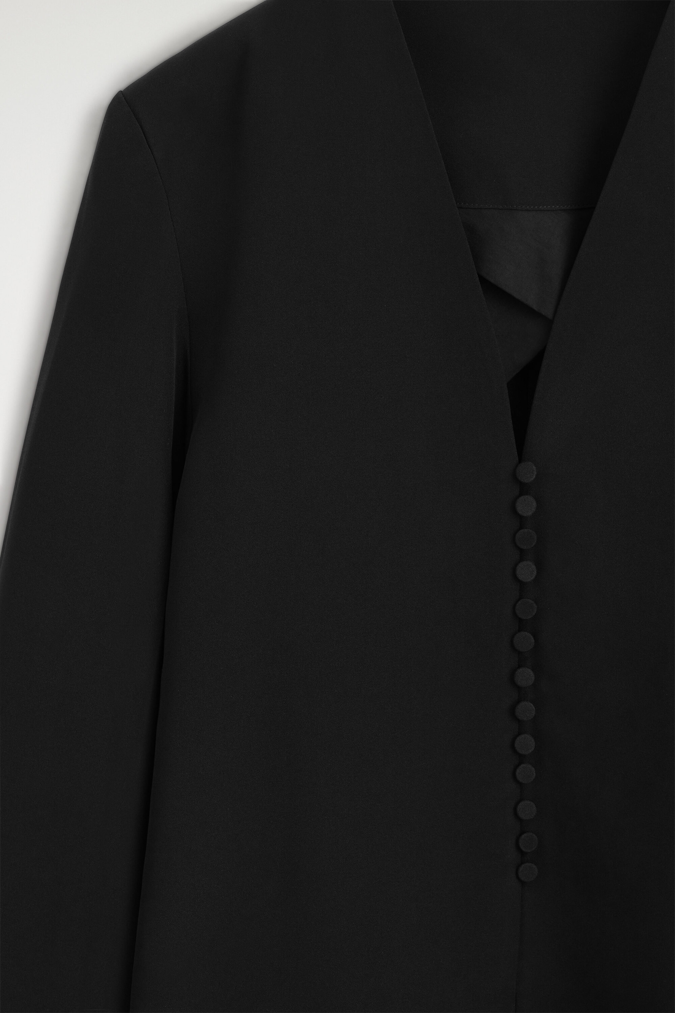 Ein schwarzer, kragenloser Blazer mit tiefem V-Ausschnitt und einer vertikalen Reihe kleiner, mit Stoff bezogener Knöpfe ist in einer Nahaufnahme von vorne zu sehen. Der glatte, matte Stoff unterstreicht die klare, minimalistische Verarbeitung des Kleidungsstücks. Ein schwarzer, kragenloser Blazer mit tiefem V-Ausschnitt und einer vertikalen Reihe kleiner, mit Stoff bezogener Knöpfe ist in einer Nahaufnahme von vorne zu sehen. Der glatte, matte Stoff unterstreicht die klare, minimalistische Verarbeitung des Kleidungsstücks.