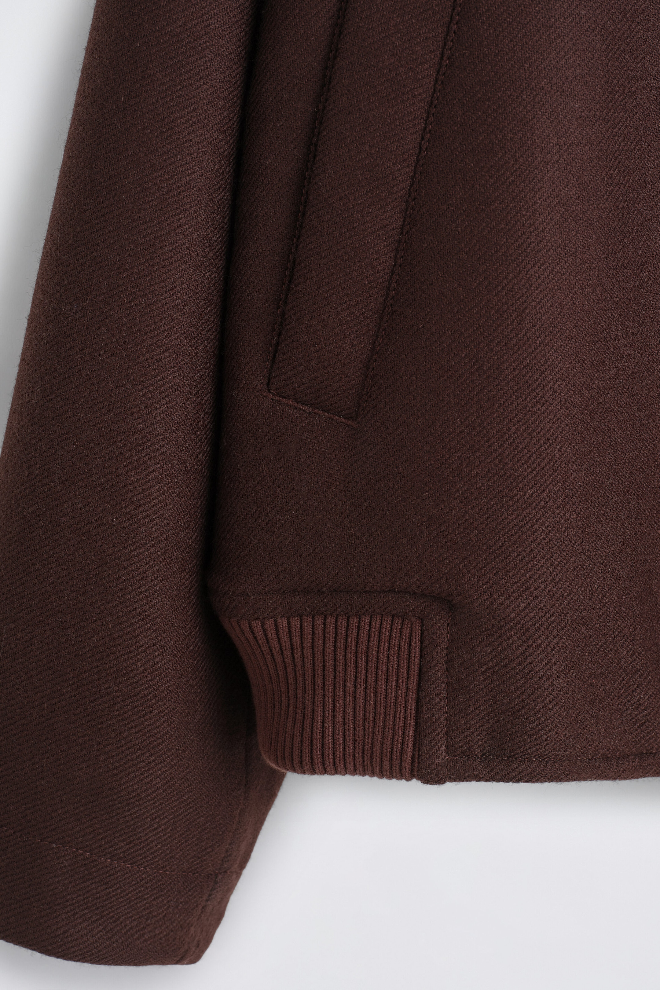 A dark brown jacket is shown in close-up detail, featuring textured twill fabric, a vertical welt pocket, and a ribbed knit hem. A dark brown jacket is shown in close-up detail, featuring textured twill fabric, a vertical welt pocket, and a ribbed knit hem.