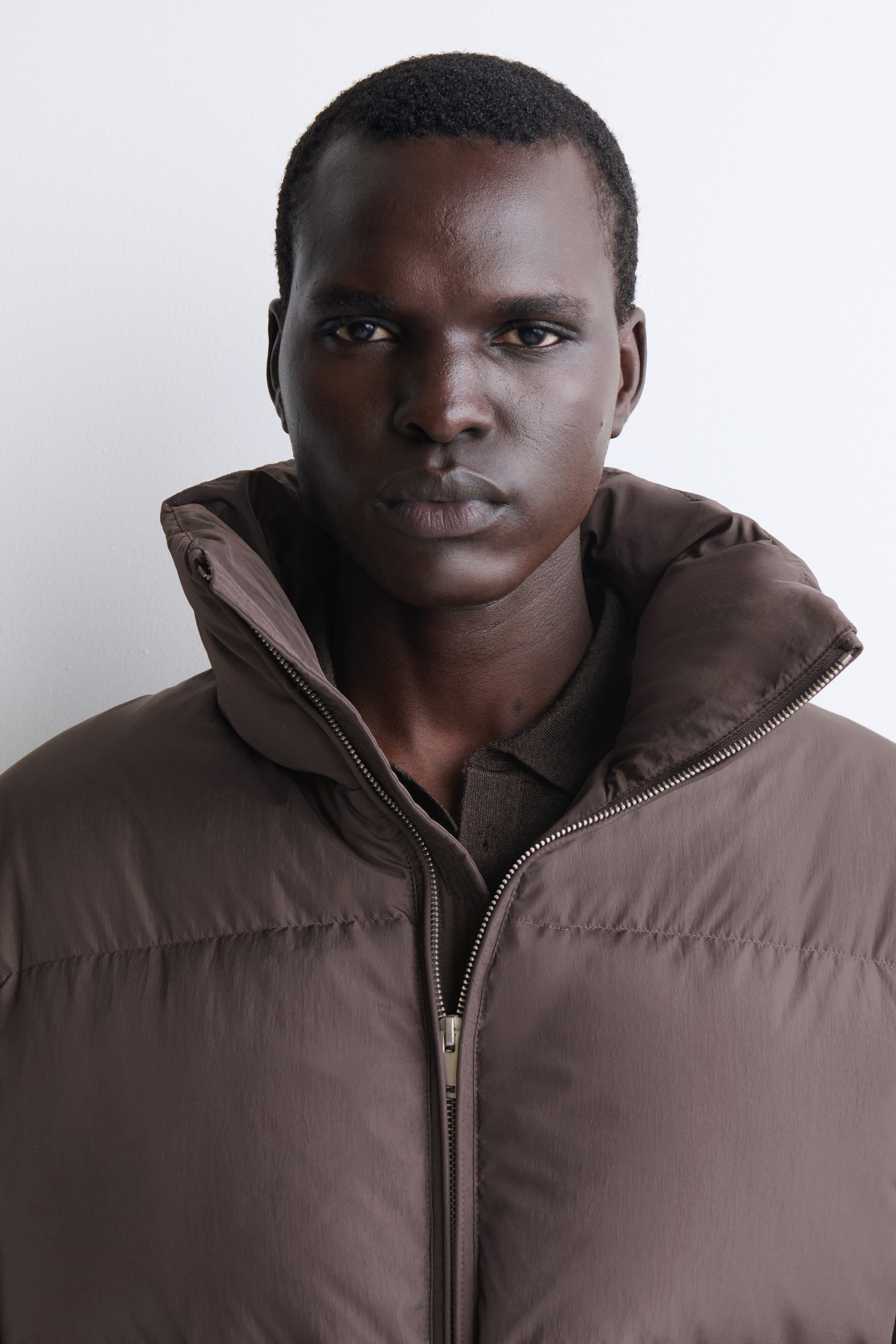 A brown funnel-neck nylon down puffer jacket is shown in a close-up front view, highlighting its high, padded collar and horizontal quilting. The jacket is worn partially unzipped, revealing a silver-tone metal zipper and a dark brown collared shirt layered underneath. A brown funnel-neck nylon down puffer jacket is shown in a close-up front view, highlighting its high, padded collar and horizontal quilting. The jacket is worn partially unzipped, revealing a silver-tone metal zipper and a dark brown collared shirt layered underneath.