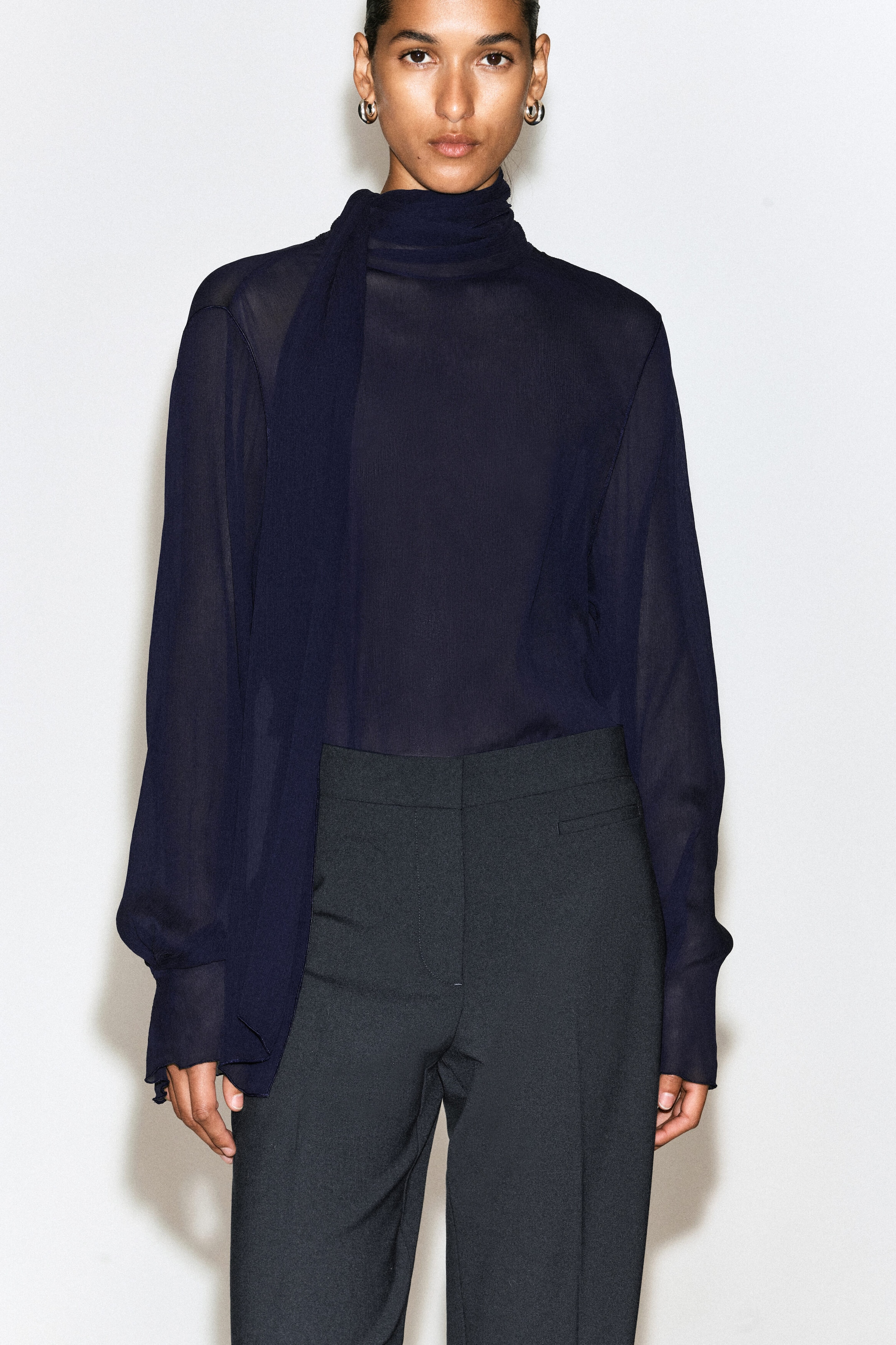 The dark grey twill cropped flared pants feature a high-rise waist with a clean, structured waistband and a single welt pocket at the side. They are shown in a tailored front view with a sharp centre crease, styled with a tucked-in sheer navy blue blouse.