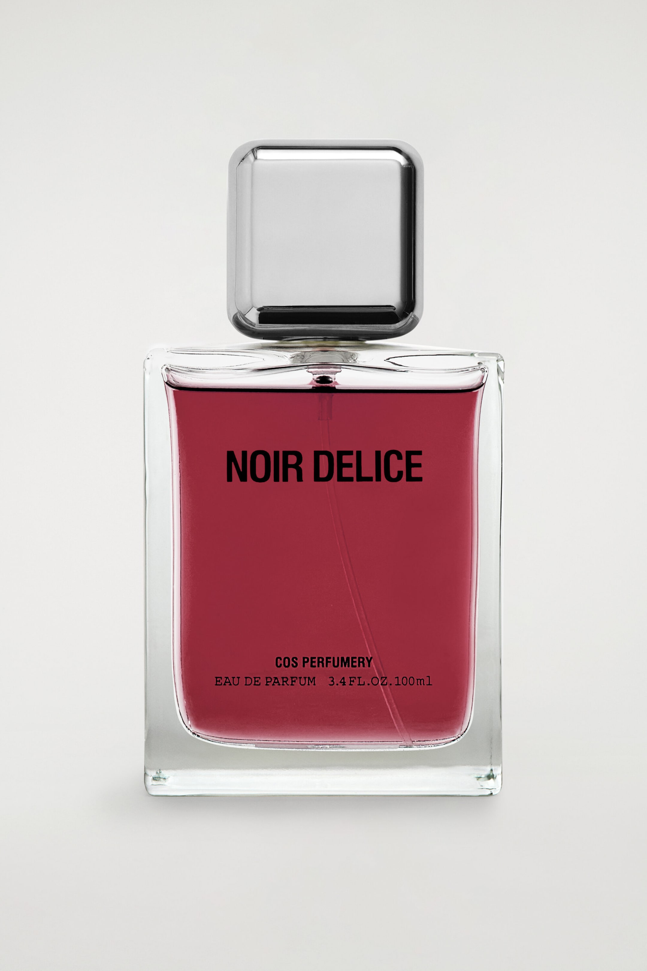 The COS PERFUMERY Eau de Parfum bottle is standing upright and facing forwards, showcasing a rectangular clear glass body filled with a dark berry-red liquid. It features a square, polished metallic silver cap and displays black printed text reading "NOIR DELICE" in the centre, with "COS PERFUMERY EAU DE PARFUM 3.4 FL.OZ. 100ml" near the bottom.