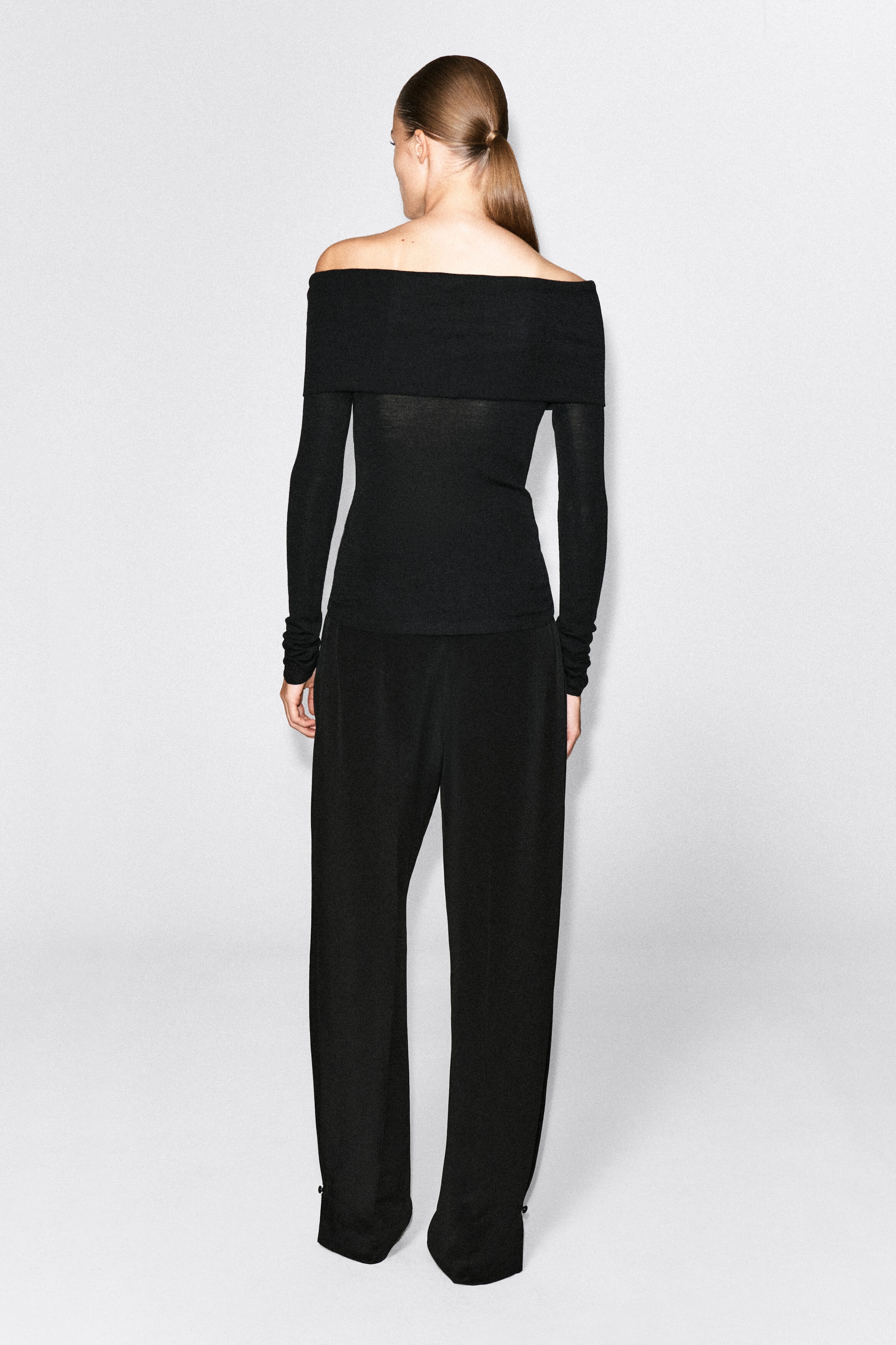 A black off-the-shoulder merino wool top is worn by the model, shown from the back. The long-sleeved garment features a wide, folded band across the shoulders and is paired with matching black trousers.
