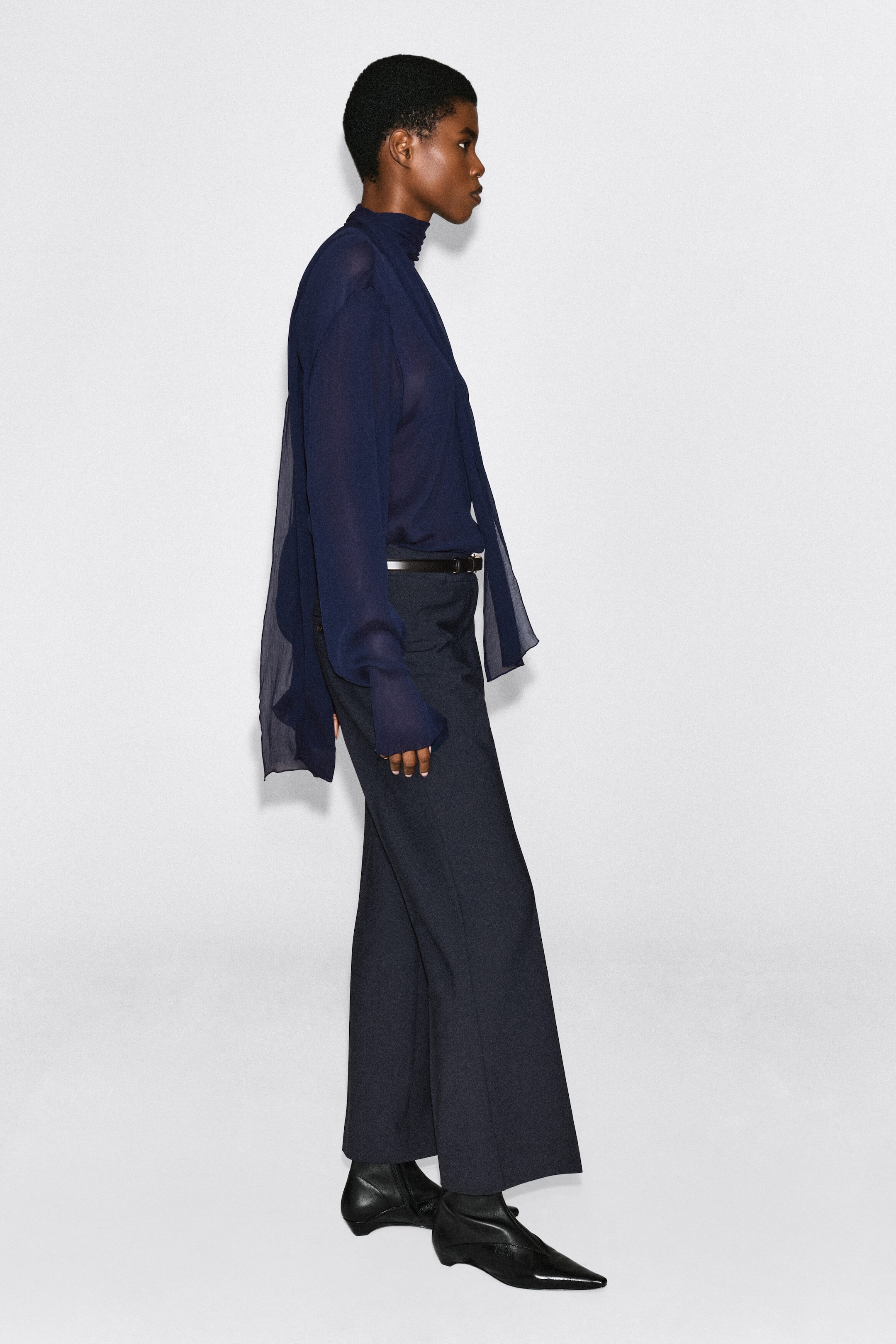 The model is wearing a dark navy blue, semi-sheer long-sleeved top, featuring a high neck with a draped scarf detail. The top is tucked into dark wide-leg trousers with a thin belt and styled with black pointed-toe boots. The model is wearing a dark navy blue, semi-sheer long-sleeved top, featuring a high neck with a draped scarf detail. The top is tucked into dark wide-leg trousers with a thin belt and styled with black pointed-toe boots.