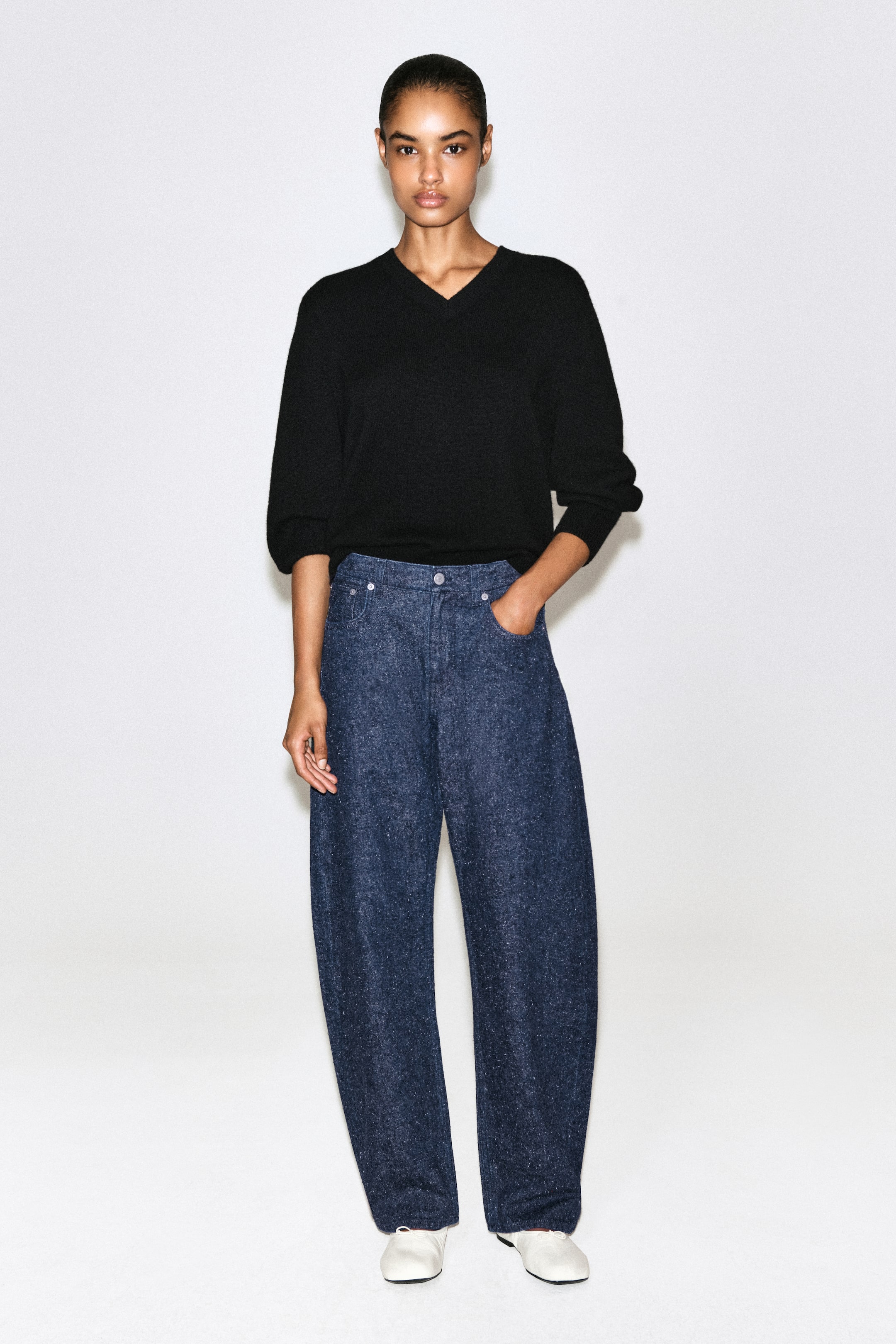 Ver imagen más grande: Estos vaqueros de cintura alta y pernera ancha, confeccionados en denim azul oscuro texturizado con efecto moteado, se muestran de frente y presentan un corte relajado y holgado, además de un diseño clásico de cinco bolsillos. Se combinan con un jersey negro de cuello en V y zapatos planos blancos, destacando su caída estructurada pero informal.