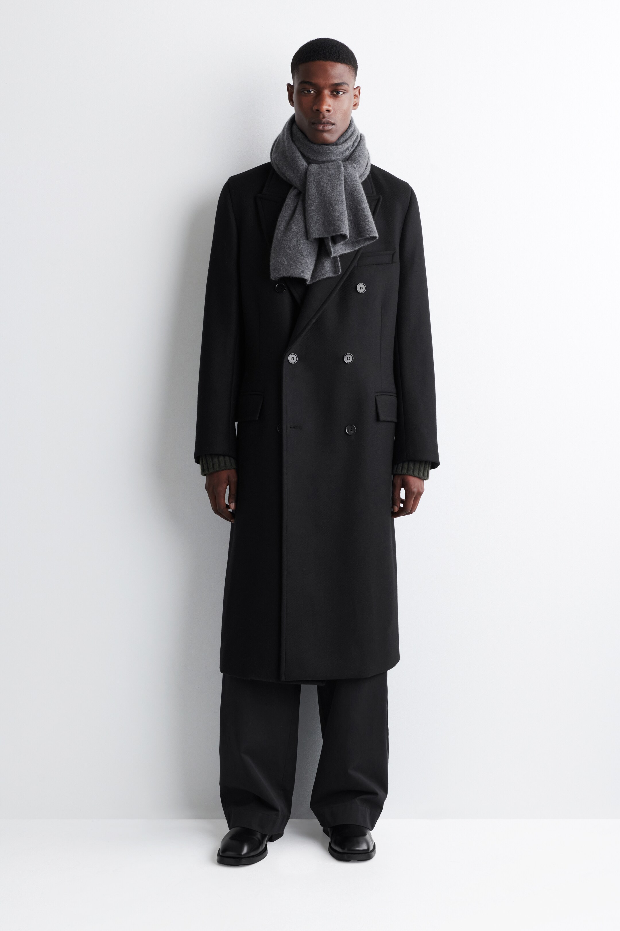 View larger image: A soft, enveloping gray scarf is being worn by the model, draped around the neck with its ends hanging down the front over a black double-breasted overcoat.