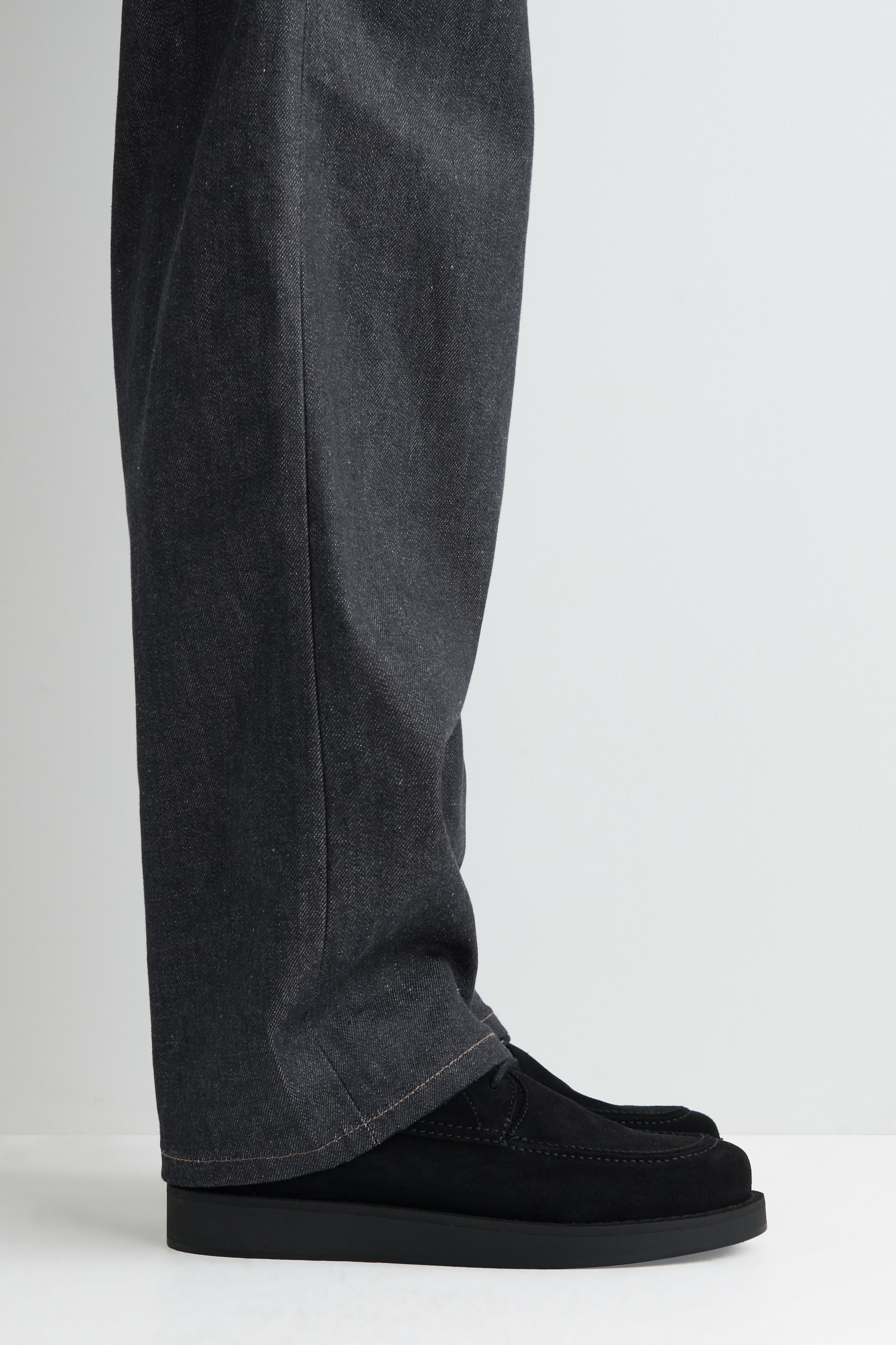 Grotere afbeelding bekijken: Donker houtskool wijde broek, met lichtbruine contrasterende stiksels bij de zoom, rusten over zwarte suède mocassin-stijl schoenen. De schoenen worden vanuit een zijhoek gepresenteerd, met een dikke, platte zwarte zool en een gestikte neus.