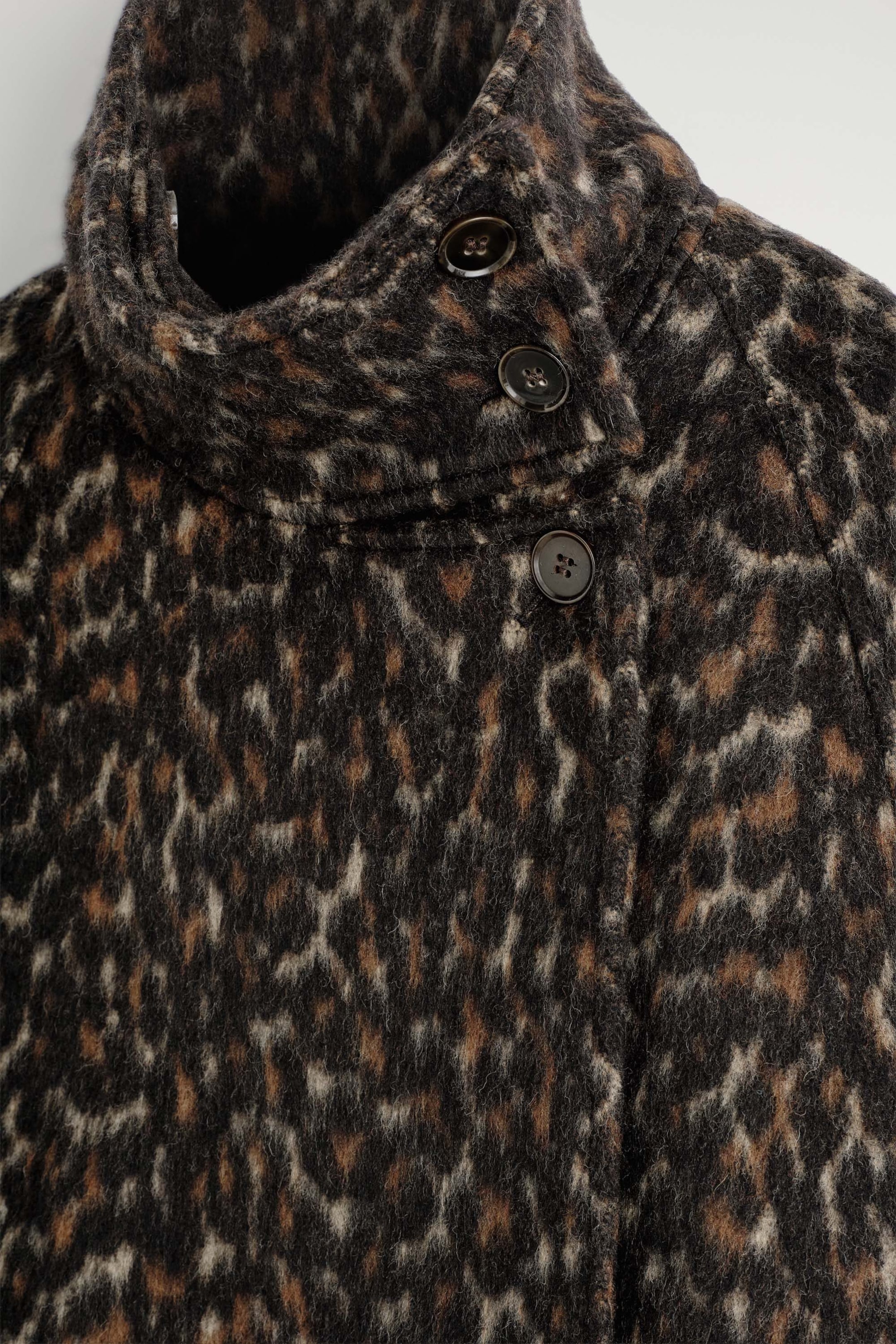 Ein Wollmix-Mantel mit Leopardenmuster und Stehkragen ist in einer Nahaufnahme von vorne zu sehen und zeigt seinen flauschigen, strukturierten Stoff. Der Mantel verfügt über einen asymmetrischen, hohen Kragen, der mit drei dunklen, runden Knöpfen verschlossen wird. Ein Wollmix-Mantel mit Leopardenmuster und Stehkragen ist in einer Nahaufnahme von vorne zu sehen und zeigt seinen flauschigen, strukturierten Stoff. Der Mantel verfügt über einen asymmetrischen, hohen Kragen, der mit drei dunklen, runden Knöpfen verschlossen wird.