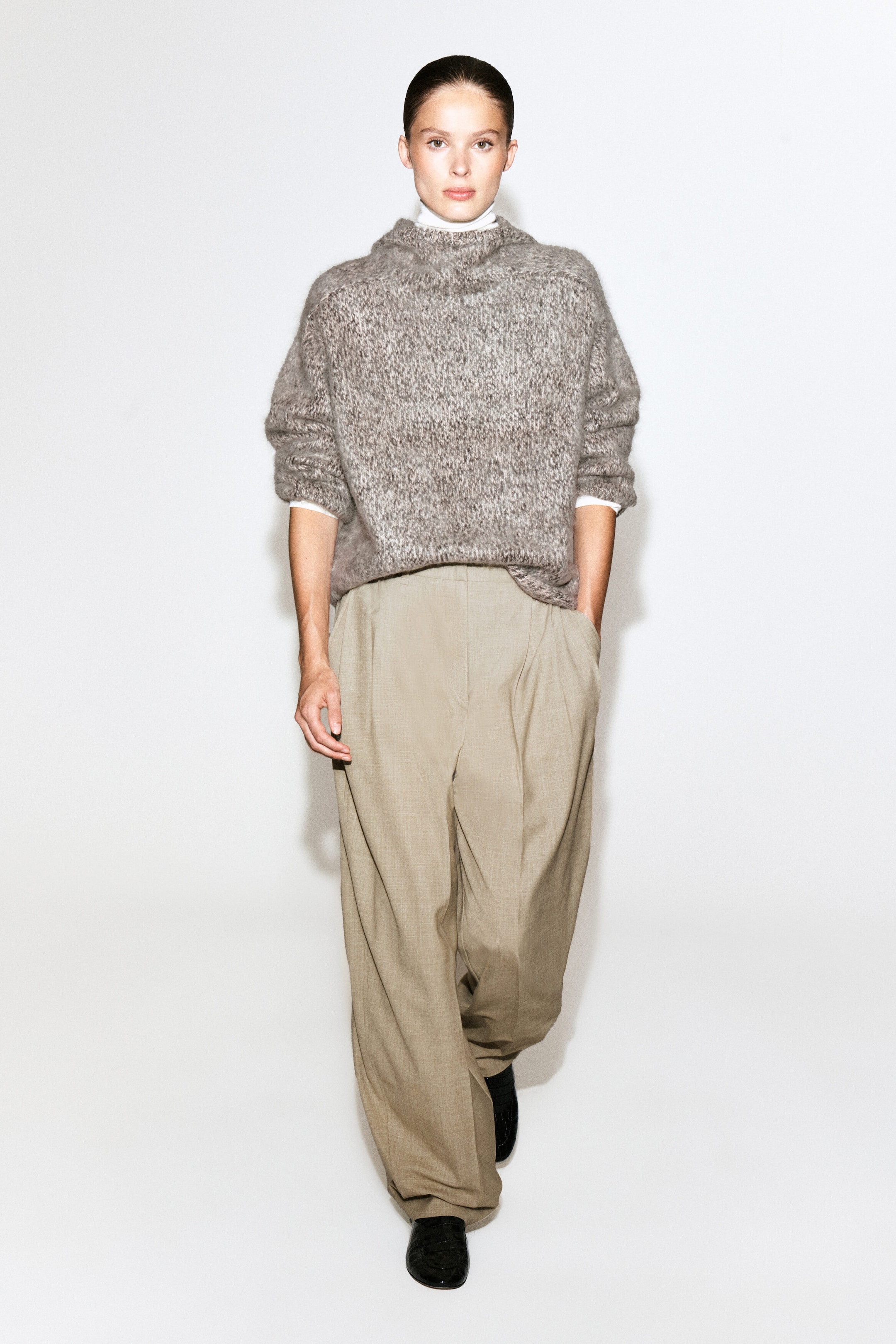 A chunky brushed-cashmere hoodie in a textured grey-brown mélange is being worn by the model over a white turtleneck top. The oversized, relaxed-fit knitwear is styled with wide-leg beige trousers. A chunky brushed-cashmere hoodie in a textured grey-brown mélange is being worn by the model over a white turtleneck top. The oversized, relaxed-fit knitwear is styled with wide-leg beige trousers.