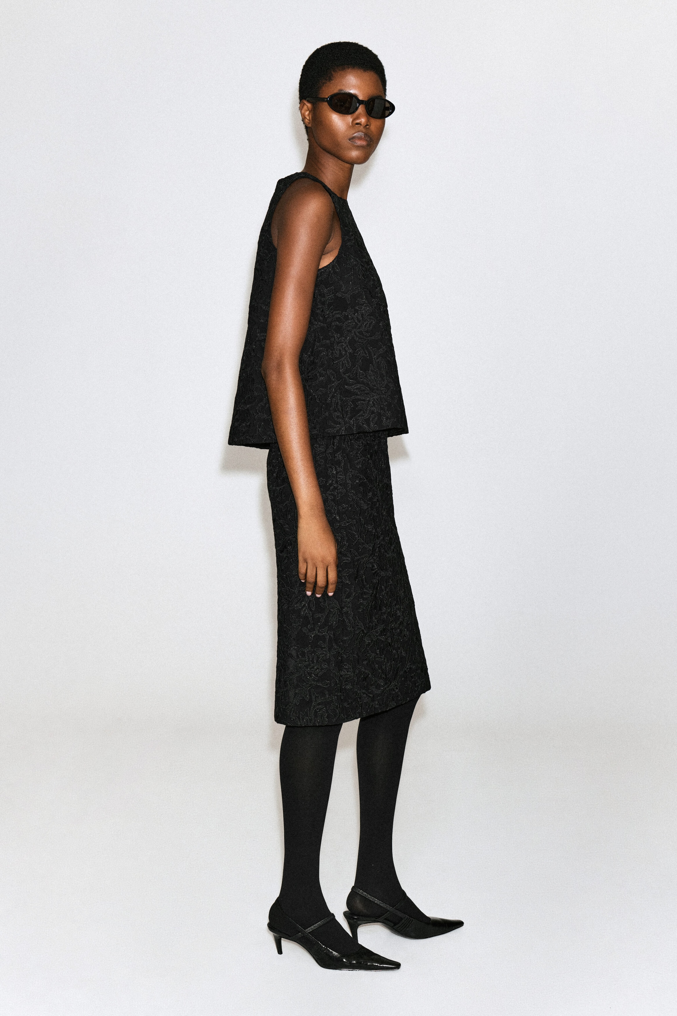 A black midi skirt featuring textured floral embroidery is being worn by the model, who is standing in side profile. The skirt is paired with a matching sleeveless top, black tights, and black pointed-toe heels. A black midi skirt featuring textured floral embroidery is being worn by the model, who is standing in side profile. The skirt is paired with a matching sleeveless top, black tights, and black pointed-toe heels.