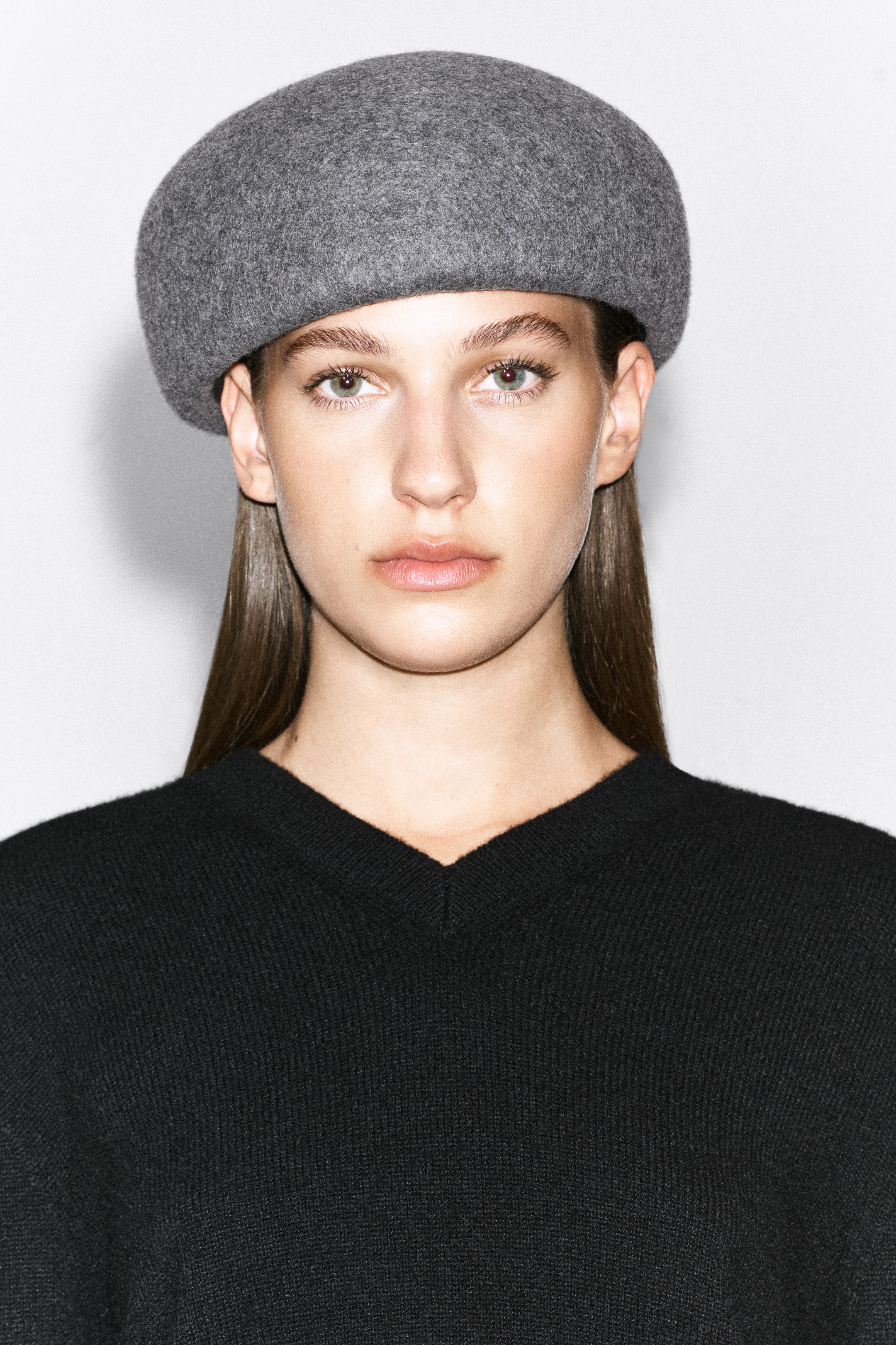 Grotere afbeelding bekijken: Een grijze, vormgegeven wollen baret met een gestructureerd, rond ontwerp wordt gedragen op het hoofd van het model. De baret is gestyled met een zwarte gebreide trui met V-hals.