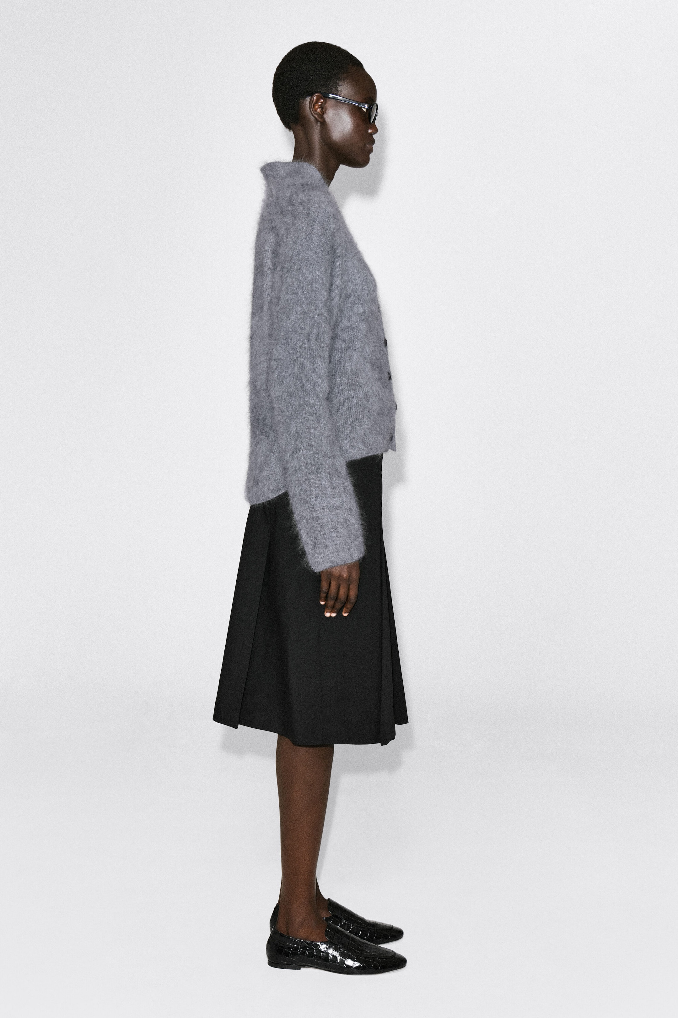 The draped brushed merino wool cardigan, facing right, showcases a fuzzy, textured knit in a gray hue with a relaxed fit. The cardigan features long sleeves and a button-front closure, draping softly at the waist over a black pleated skirt.