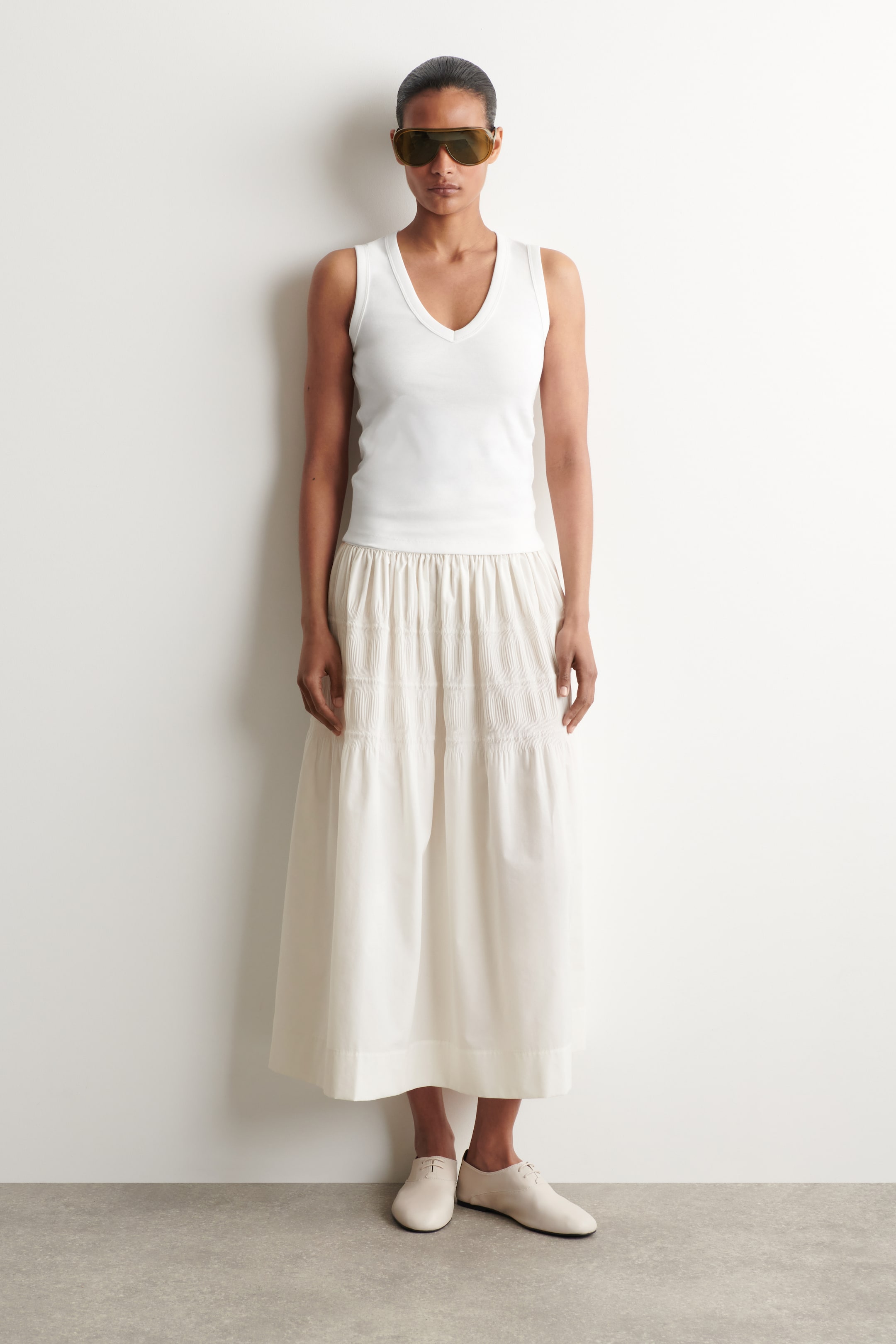 Agrandir l'image: La mannequin porte une jupe midi blanc cassé, assortie d'un haut blanc sans manches. La jupe présente une taille élastiquée froncée et une construction à étages avec des détails de smocks horizontaux, tombant jusqu'au milieu du mollet.