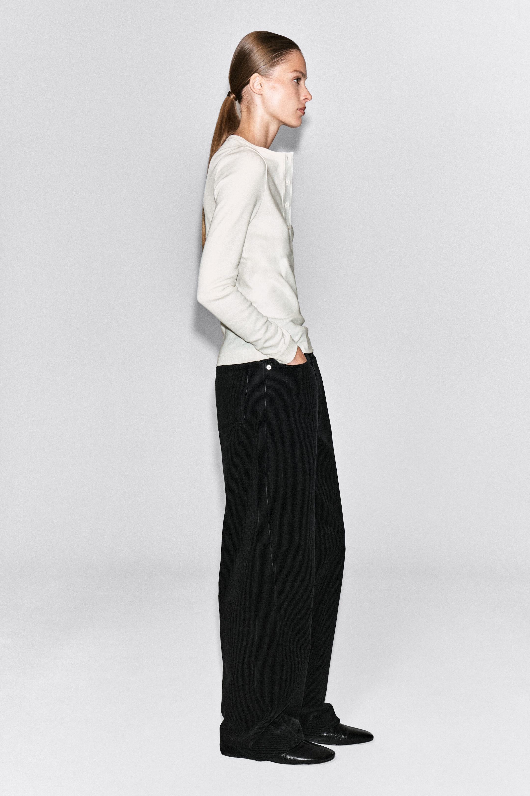 A ribbed cotton Henley top in off-white is worn by the model, featuring a fitted silhouette, long sleeves, and a button placket. The top is styled with wide-leg black trousers, with the model standing in profile. A ribbed cotton Henley top in off-white is worn by the model, featuring a fitted silhouette, long sleeves, and a button placket. The top is styled with wide-leg black trousers, with the model standing in profile.