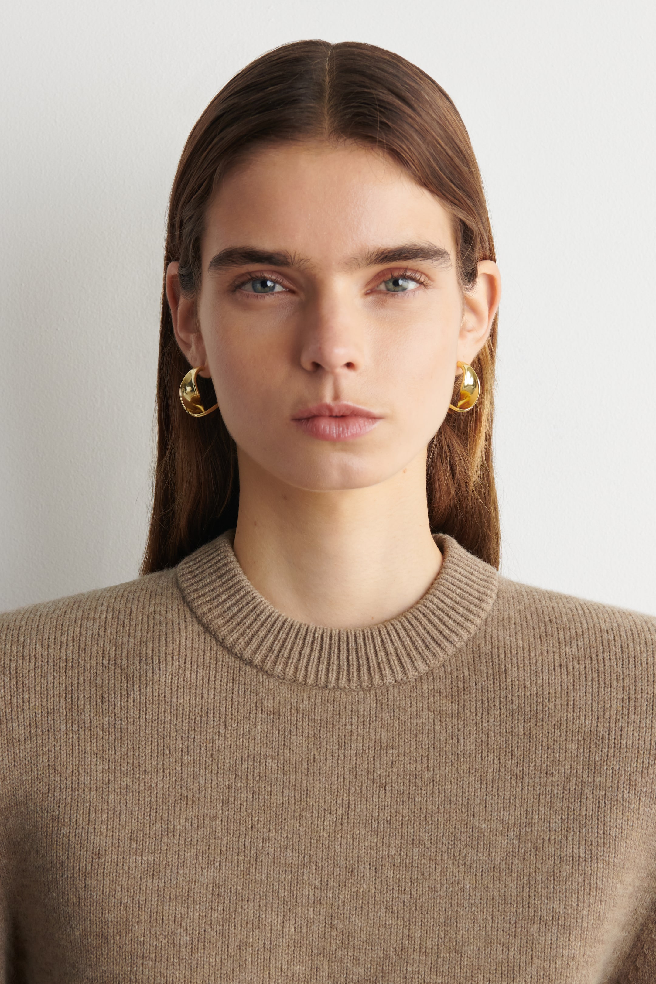View larger image: A brown crew neck jumper featuring a ribbed collar and a fine knit texture, facing front. Gold-toned, irregularly shaped hoop earrings curve around the ears, facing slightly to the side.