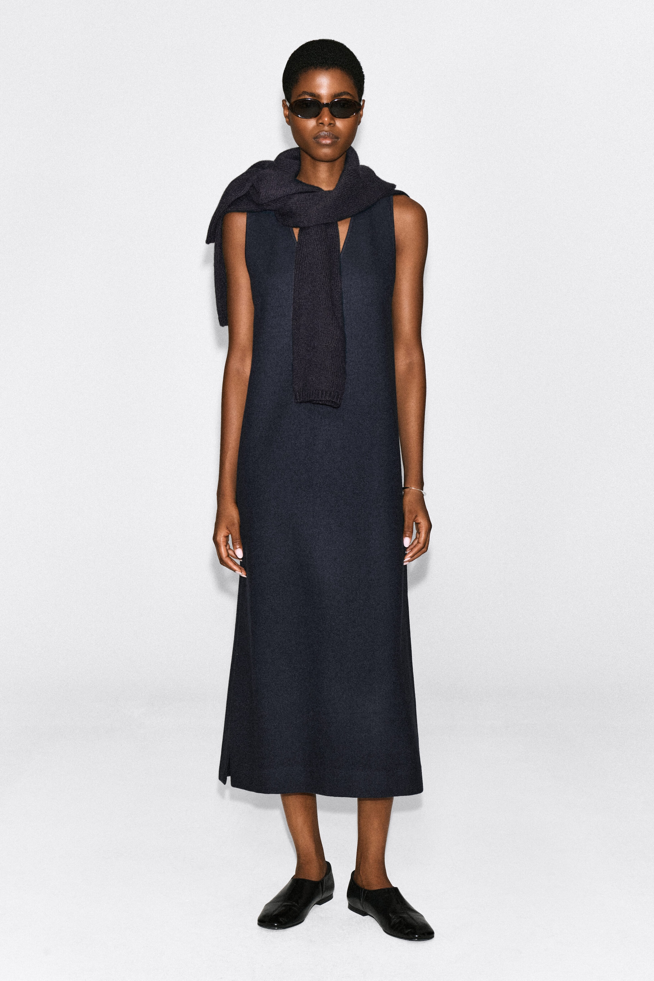 The model is wearing a navy boiled wool-blend V-neck midi dress with a sleeveless, straight silhouette. The dress is styled with a matching dark knit scarf draped over the shoulders, black sunglasses, and black flat shoes. The model is wearing a navy boiled wool-blend V-neck midi dress with a sleeveless, straight silhouette. The dress is styled with a matching dark knit scarf draped over the shoulders, black sunglasses, and black flat shoes.