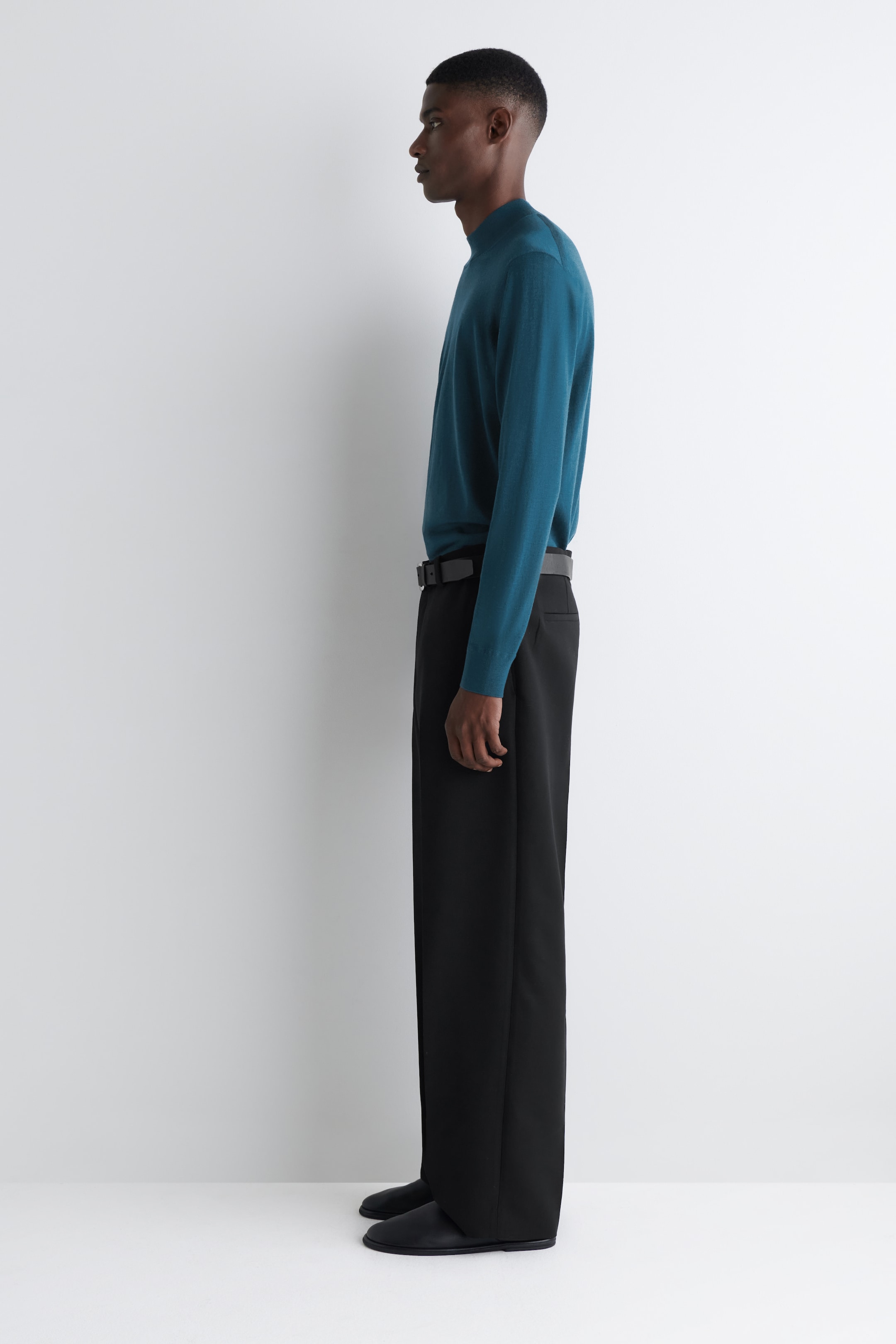 View larger image: A person is standing in left profile, wearing a teal mock-neck sweater, black wide-leg trousers with a black belt, and black pointed-toe flat shoes.
