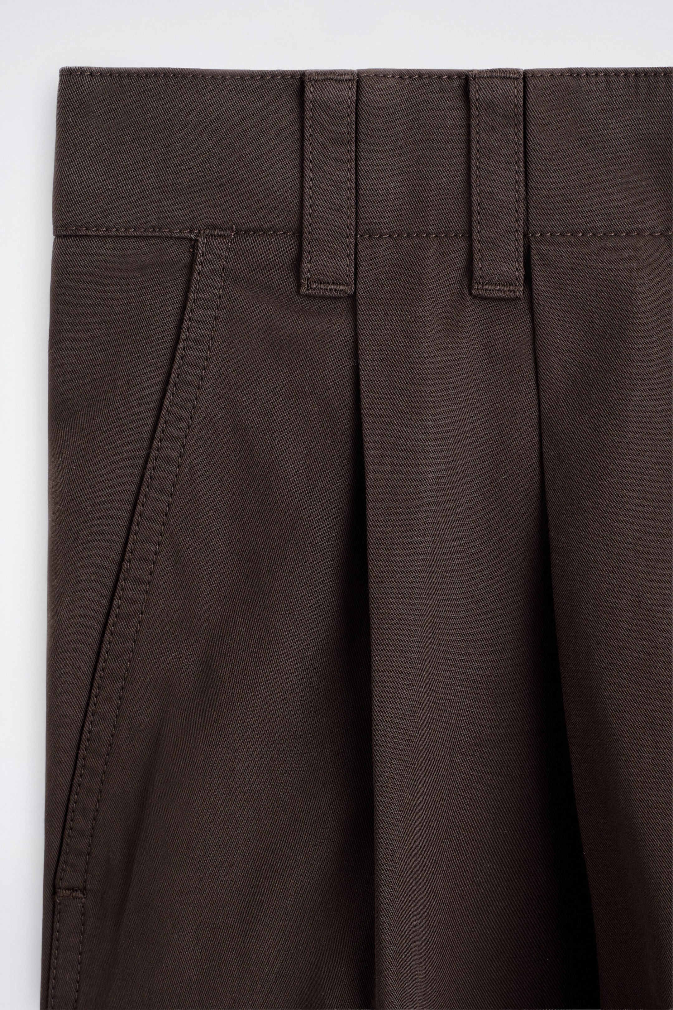 The tailored pleated cotton shorts are shown in a close-up front view, highlighting their dark brown twill construction, a structured waistband with belt loops, a single front pleat, and a slanted side pocket with visible topstitching. The tailored pleated cotton shorts are shown in a close-up front view, highlighting their dark brown twill construction, a structured waistband with belt loops, a single front pleat, and a slanted side pocket with visible topstitching.
