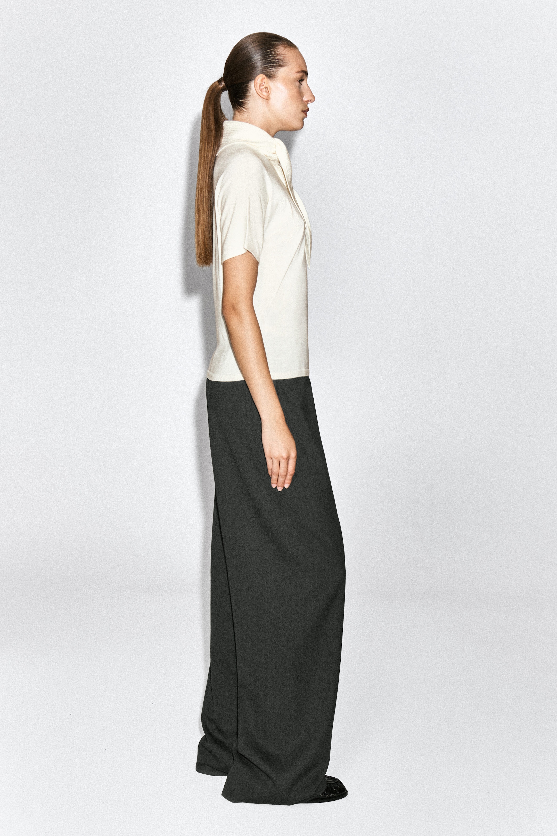 A cream-colored, scarf-detail merino wool-blend T-shirt is worn by the model, featuring short sleeves and an integrated draped collar detail at the neckline. The T-shirt is styled with dark, wide-leg trousers.