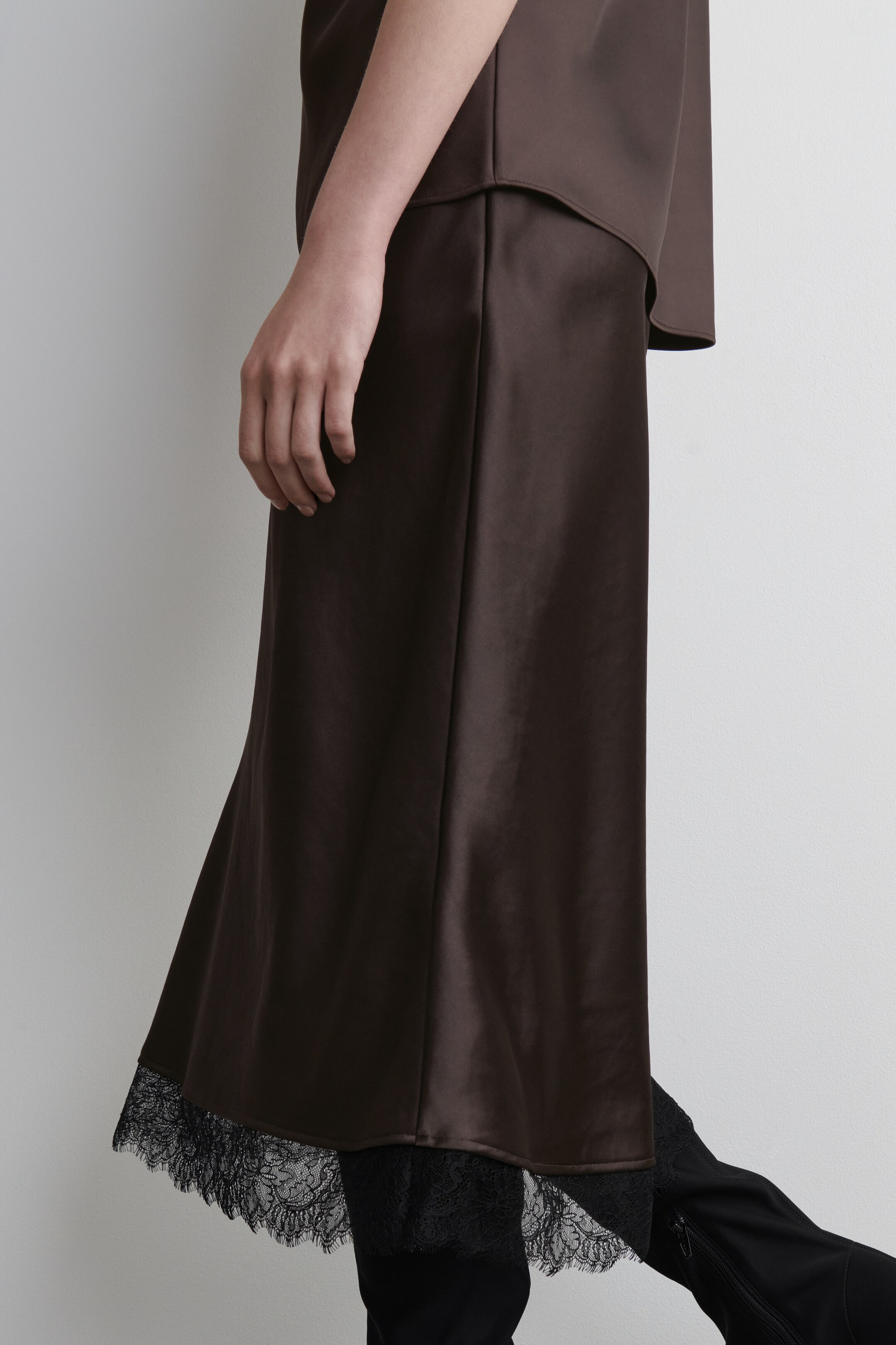 View larger image: A dark brown satin skirt is flowing, with a black lace trim visibly peeking out from its hem. The skirt is being styled with a matching dark brown top covering its waistband and is layered over black pants or boots.
