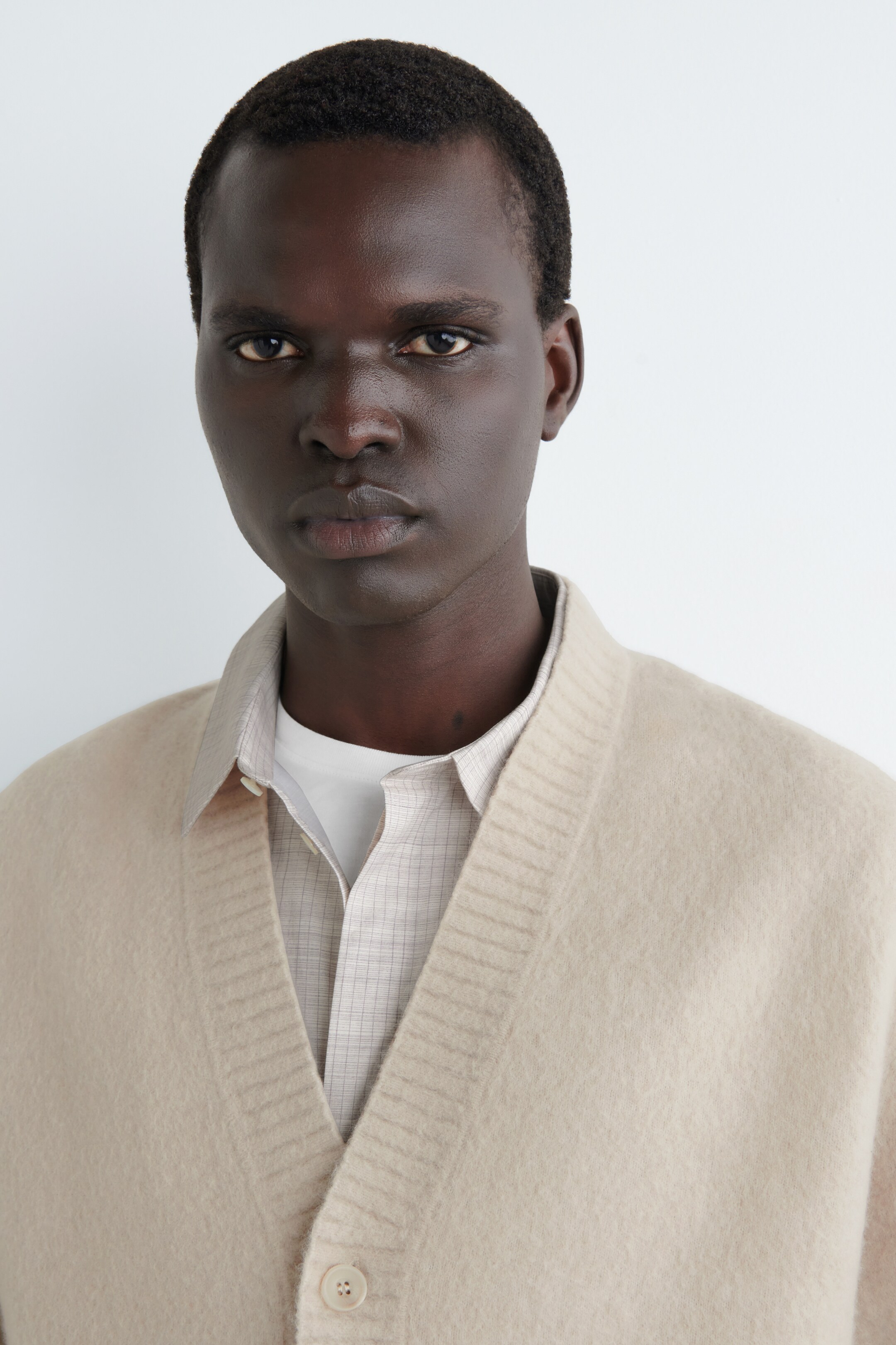 Ver imagen más grande: Se presenta un cárdigan beige de cara, que presenta una textura suave y cepillada y una abertura en V con canalé a lo largo de los bordes. Está superpuesto sobre una camisa de cuello de color claro con un discreto estampado de cuadros, que a su vez está sobre una camiseta blanca, y un solo botón a juego es visible en el frente abierto.