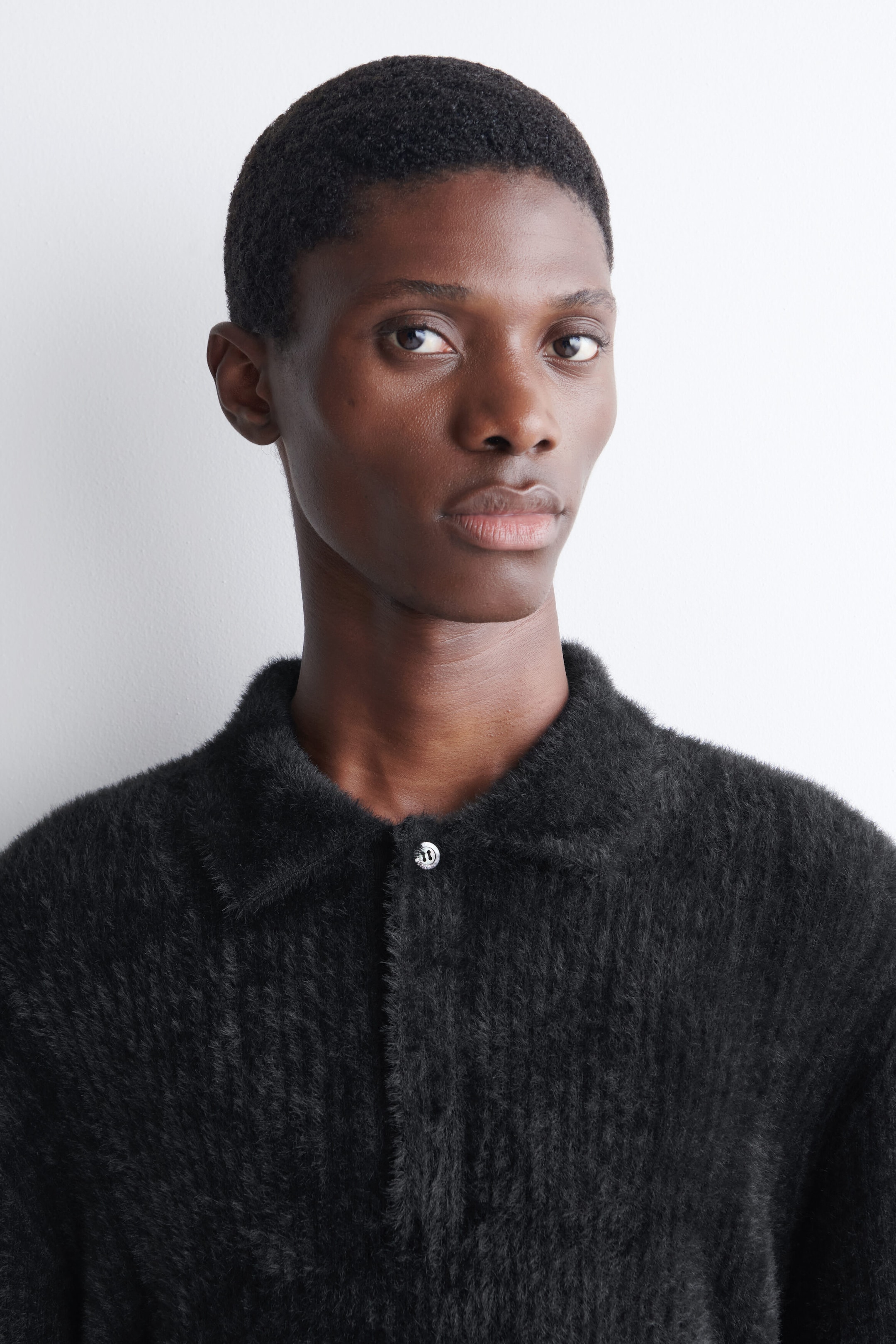 The model is wearing a dark gray knitted polo shirt with a highly textured, brushed finish. The shirt features a classic flat collar and a single-button closure at the top of the placket. The model is wearing a dark gray knitted polo shirt with a highly textured, brushed finish. The shirt features a classic flat collar and a single-button closure at the top of the placket.