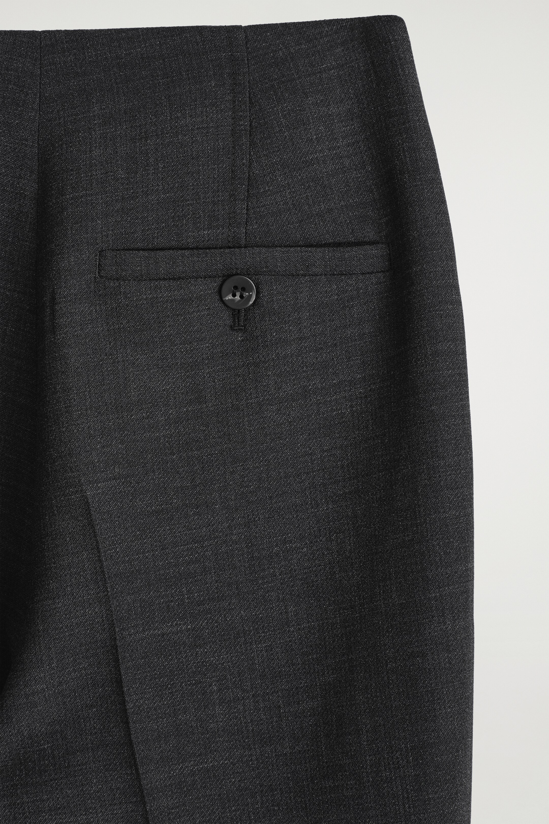 Größeres Bild anzeigen: Die dunkelgraue Hose zeigt eine Nahaufnahme einer einzelnen hinteren Leistentasche, die mit einem dunklen Vierlochknopf befestigt ist. Der Stoff weist ein feines, dezent strukturiertes Webmuster auf.