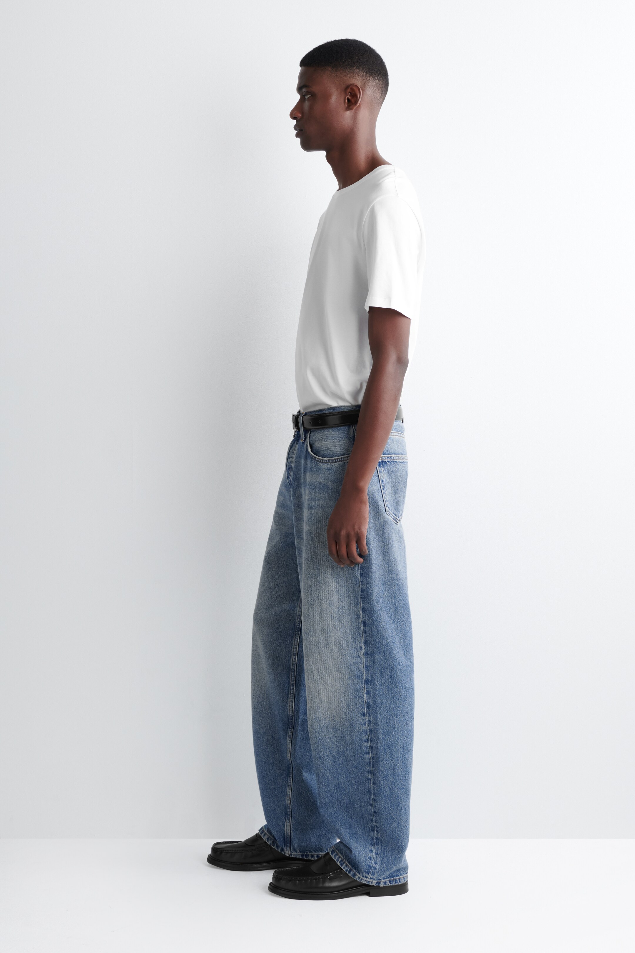Visa större bild: En vit kortärmad t-shirt med rund hals bärs, instoppad i ljusblå vida jeans som hålls uppe av ett svart bälte. Jeansen har en avslappnad passform genom benen och samlas något vid anklarna över svarta slip-on-skor med synliga tåstygn.