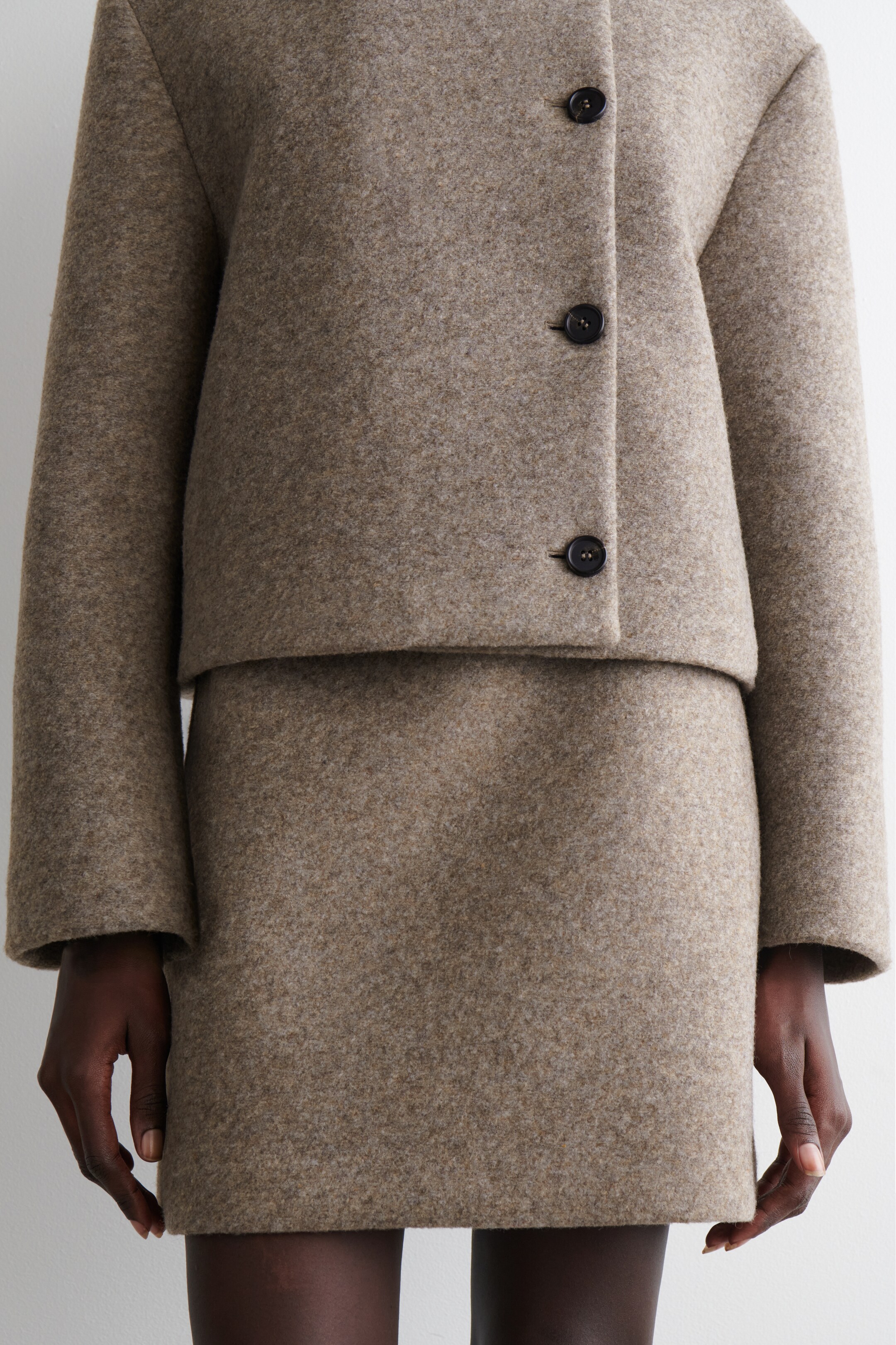 Visualizza immagine più grande: Un completo a due pezzi di colore marrone chiaro e con una texture particolare viene presentato, composto da una giacca corta con chiusura frontale a tre bottoni e maniche lunghe, abbinata a una minigonna coordinata, il tutto caratterizzato da una silhouette strutturata e squadrata.