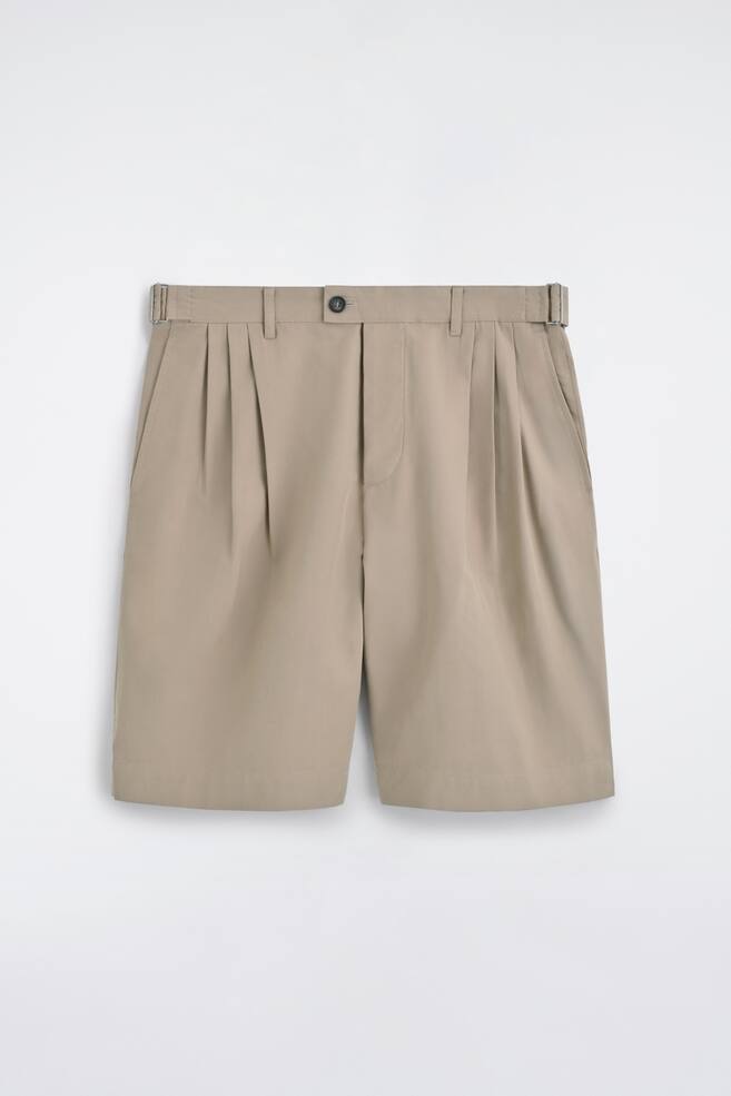 Les shorts beiges sont présentés de face, avec des plis doubles à l'avant et une fermeture à bouton à la taille. Des ajusteurs latéraux avec des boucles métalliques rectangulaires ornent la ceinture, et des poches fendues sur les côtés sont visibles. - 1