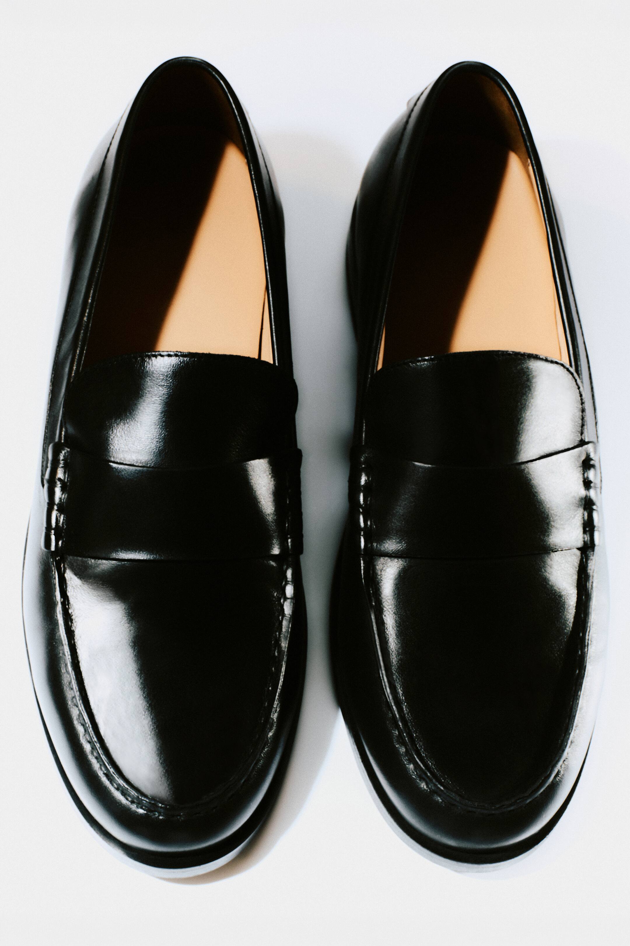 Grotere afbeelding bekijken: Een paar zwarte leren loafers worden gepresenteerd vanuit een recht van boven genomen, frontale hoek. Ze hebben een glanzende afwerking, een klassiek pennybandje over de wreef en ton-sur-ton stiksel rond de neuskap. Een lichtbruine voering is zichtbaar langs de binnenkraag.