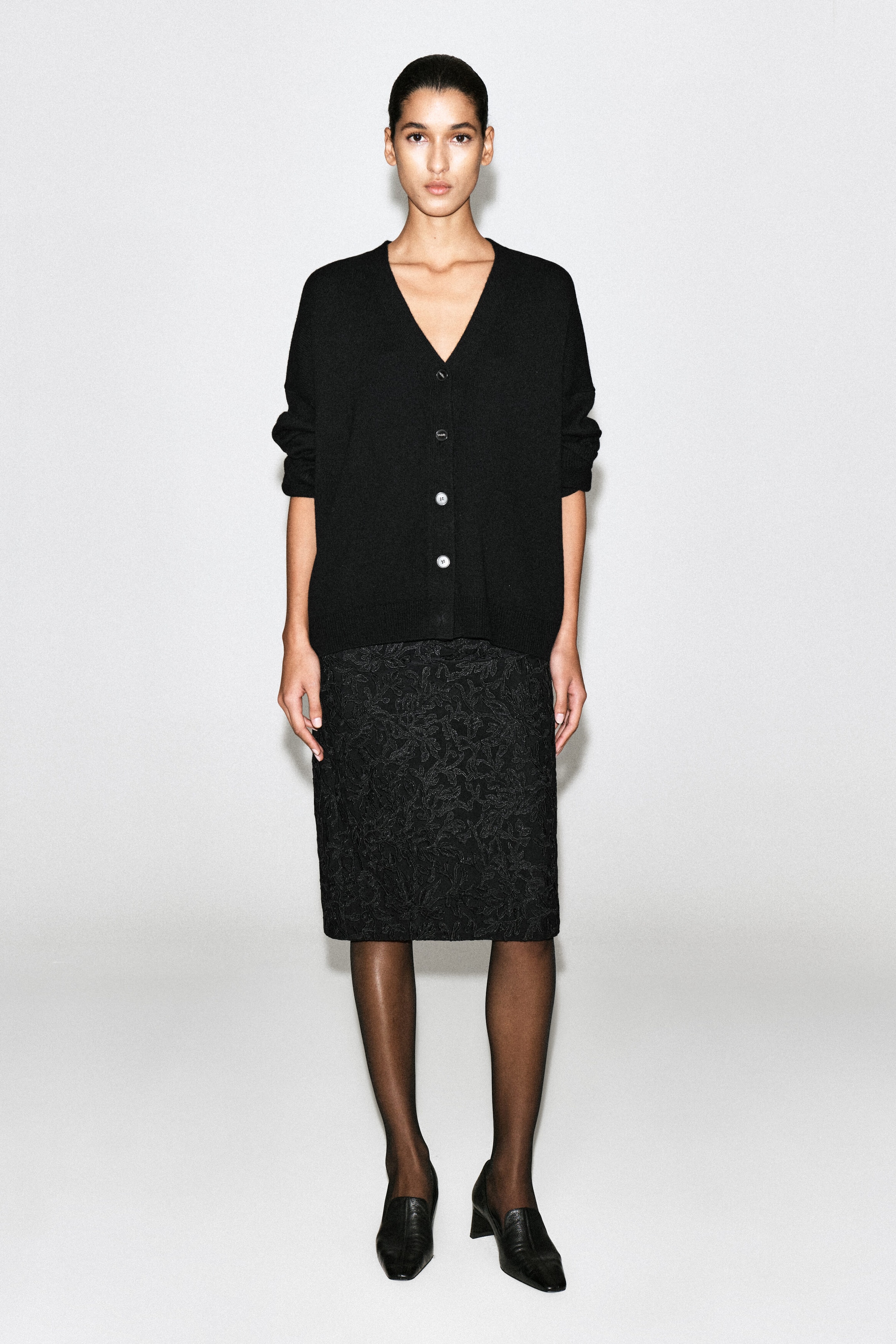 Una modella indossa un cardigan nero con scollo a V e vestibilità rilassata, ripresa frontalmente, che mette in risalto la chiusura a quattro bottoni, le spalle scese e le finiture a coste. Il cardigan è abbinato a una gonna nera al ginocchio con texture, collant neri velati e mocassini neri.